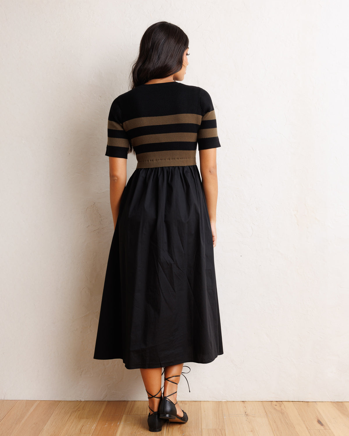 A woman is shown from behind wearing a black midi dress with brown horizontal stripes on the bodice and sleeves, paired with black lace-up heels, standing against a textured off-white wall on a light wood floor.