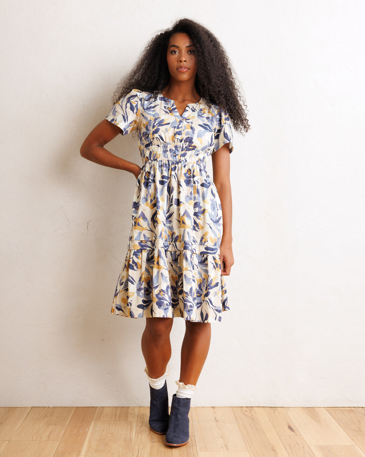 A woman modeling a short-sleeve tiered midi dress with a blue and yellow floral print on a cream background, paired with white socks and navy boots, standing against a white wall.