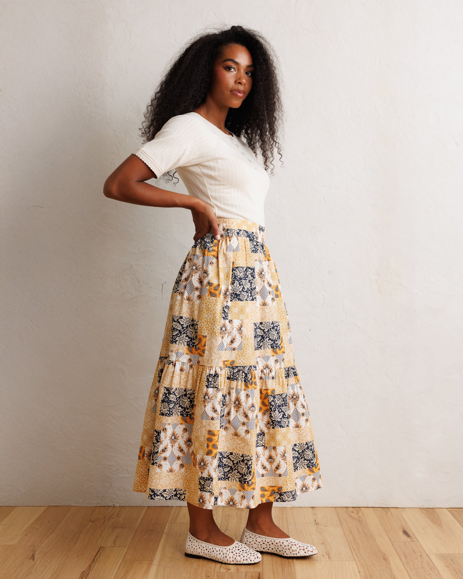 A woman stands in a studio wearing a white short-sleeve top and a long tiered skirt with a patchwork-style print in beige, mustard yellow, navy, and white. She is posed against a light textured wall on a wood floor.