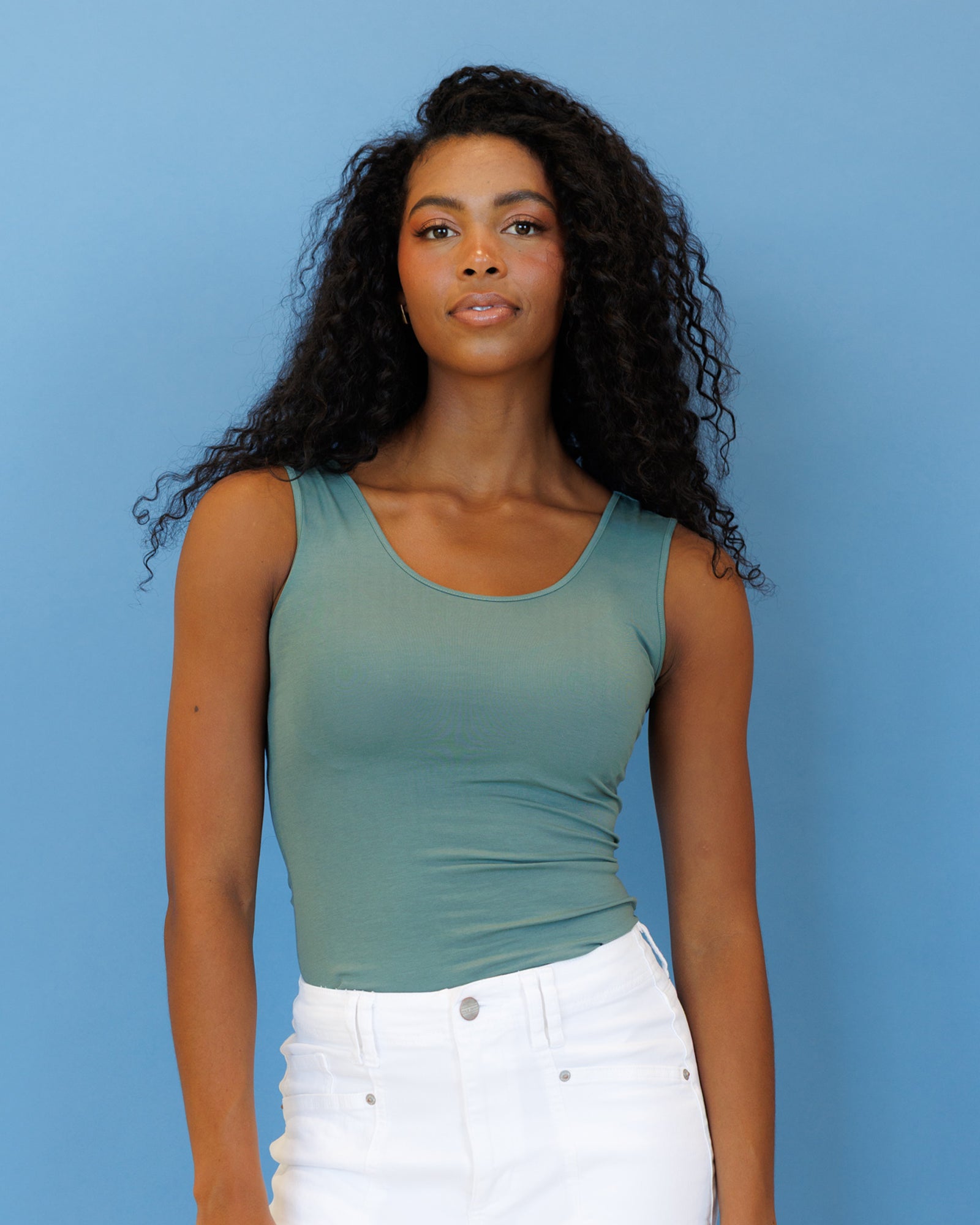 A woman with long, curly black hair wearing a fitted sage green scoop-neck tank top and white jeans, standing against a solid blue background.