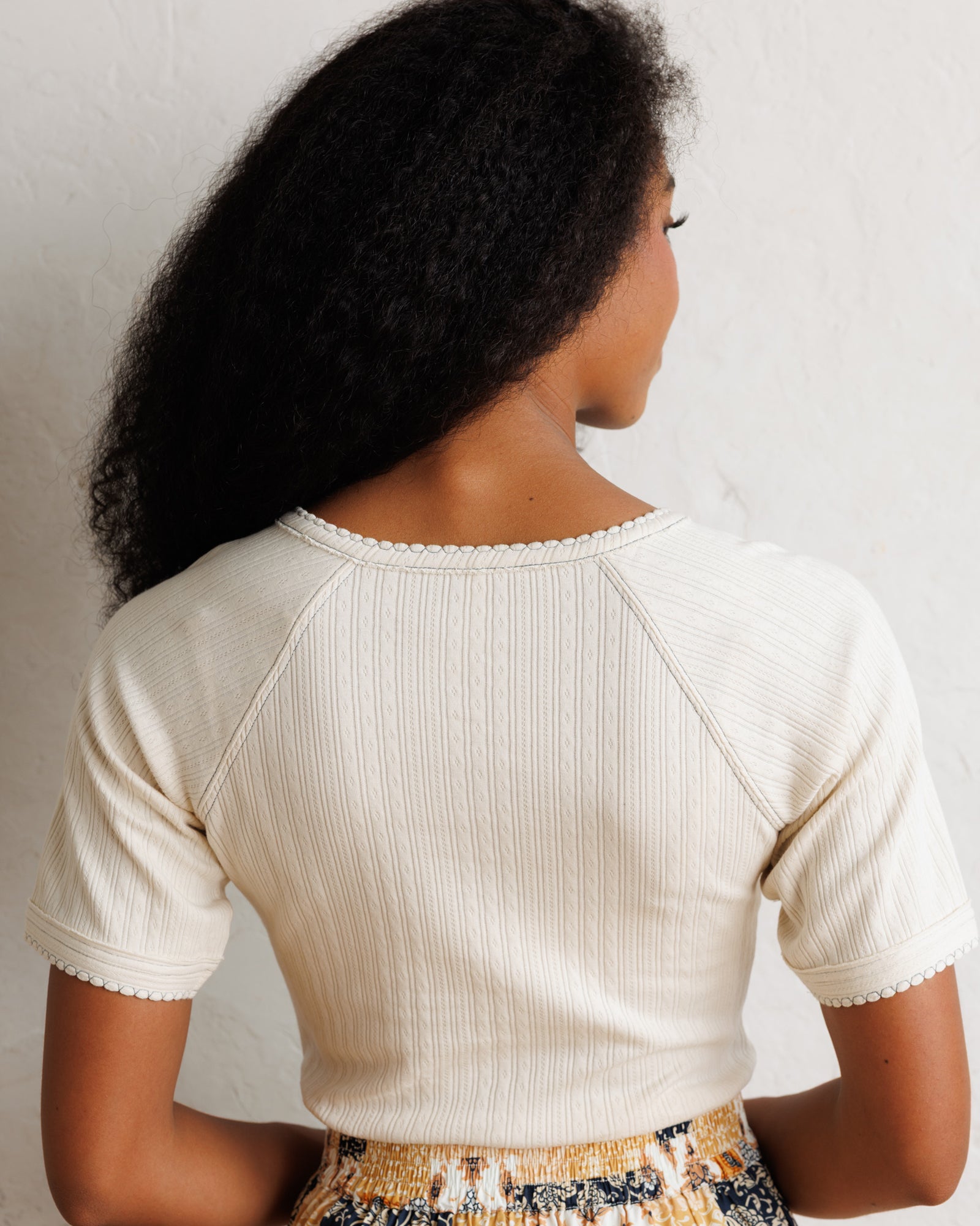 A woman shown from behind wearing a cream-colored ribbed short-sleeve crop top with scalloped edges, paired with a patterned skirt, against a textured off-white wall.