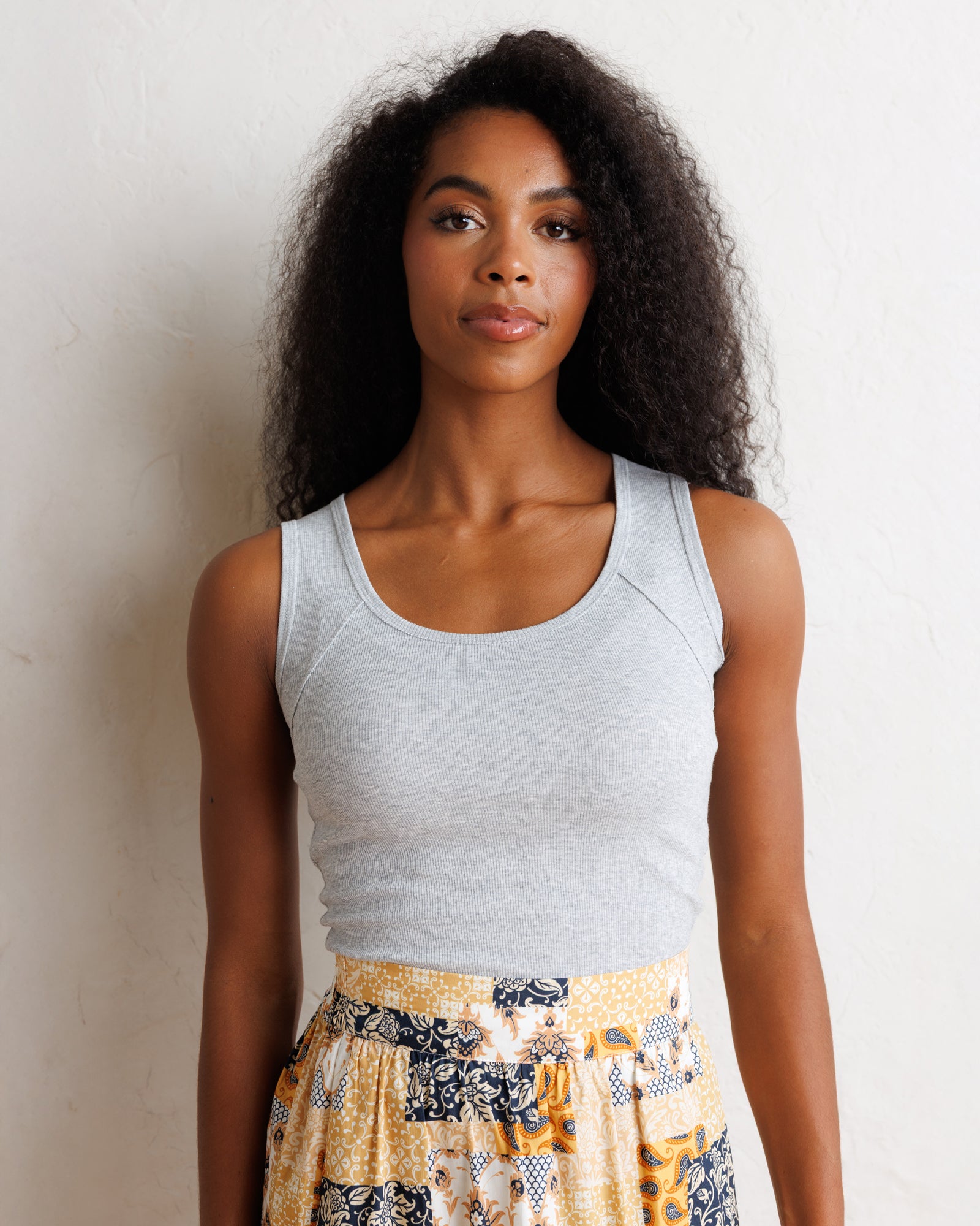 A woman with long, curly black hair is shown from the waist up, wearing a light gray sleeveless top and a patterned skirt with yellow, black, and white tones. She is posing against a textured off-white wall.