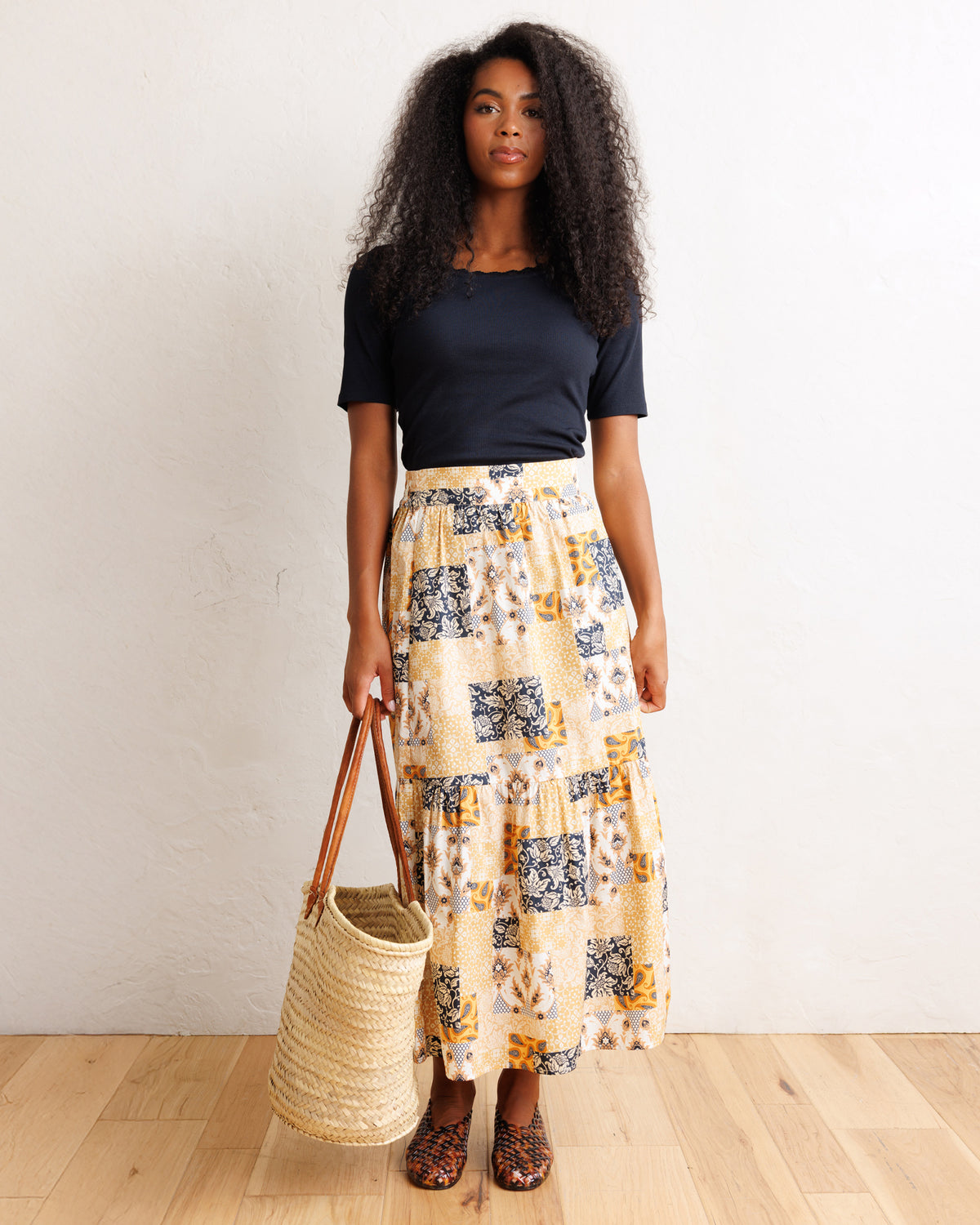 A woman stands in a room with a white wall and light wood floor, wearing a navy blue short-sleeve top and a long cream skirt with a blue and yellow abstract pattern. She holds a large woven straw tote bag.