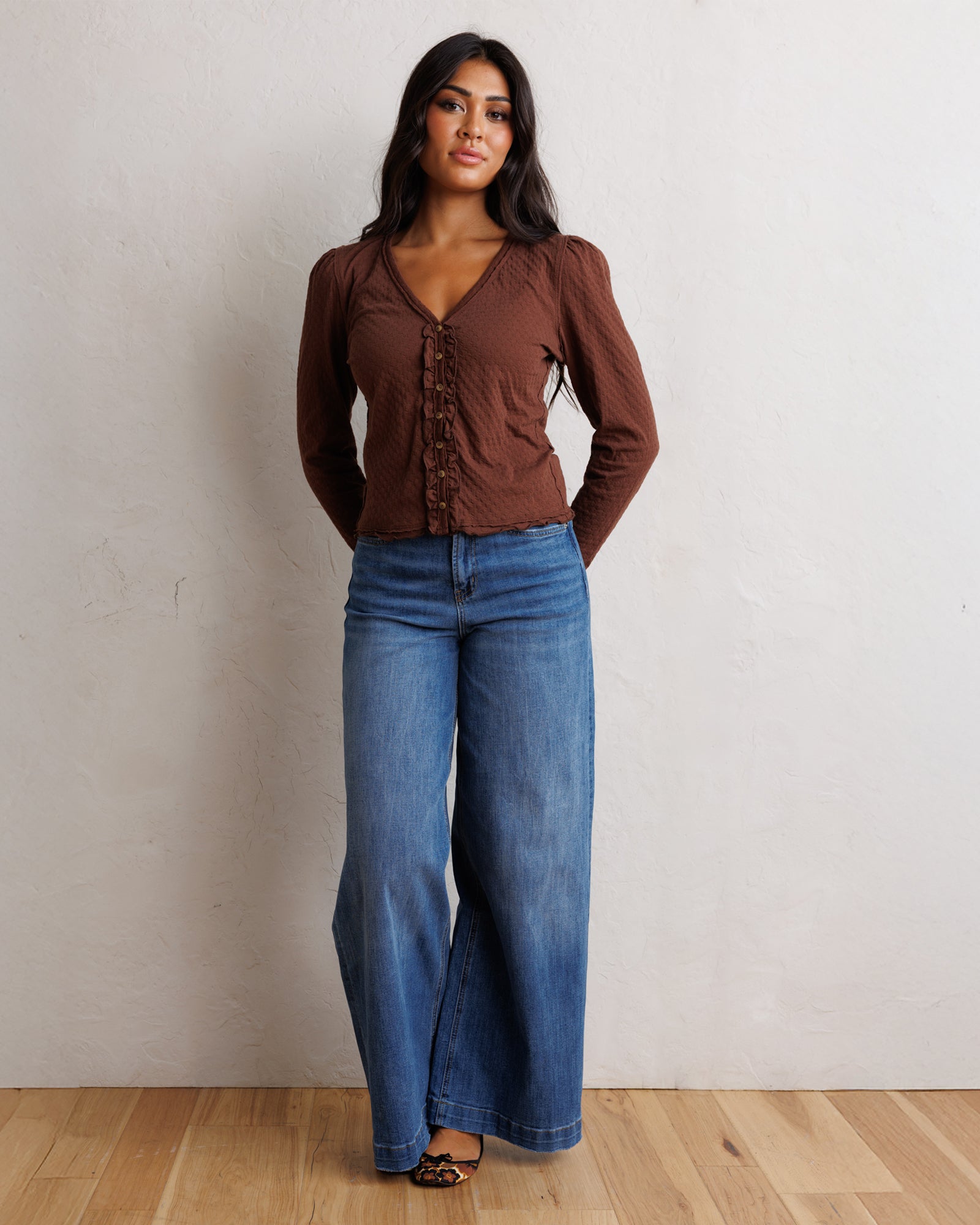 A woman stands against a textured off-white wall on a light wood floor, wearing a brown long-sleeve button-front top with gathered ruffle details down the front and wide-leg blue jeans.