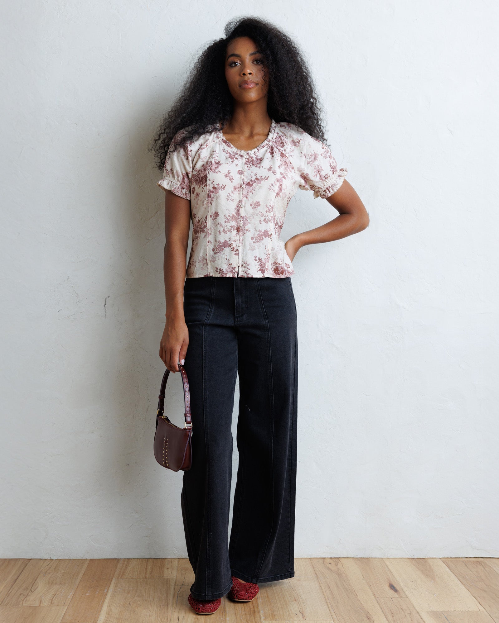 A woman stands against a white wall wearing a cream short-sleeve blouse with a dusty pink floral print, dark wide-leg corduroy pants, and red shoes, while holding a small brown handbag.