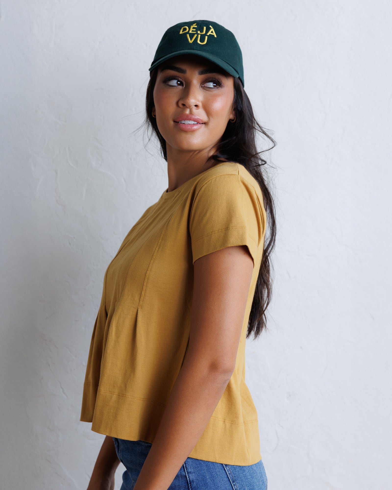 A woman wearing a mustard yellow short-sleeve top and a dark green baseball cap with yellow 'DEJA Vu' text, standing against a light gray textured wall.