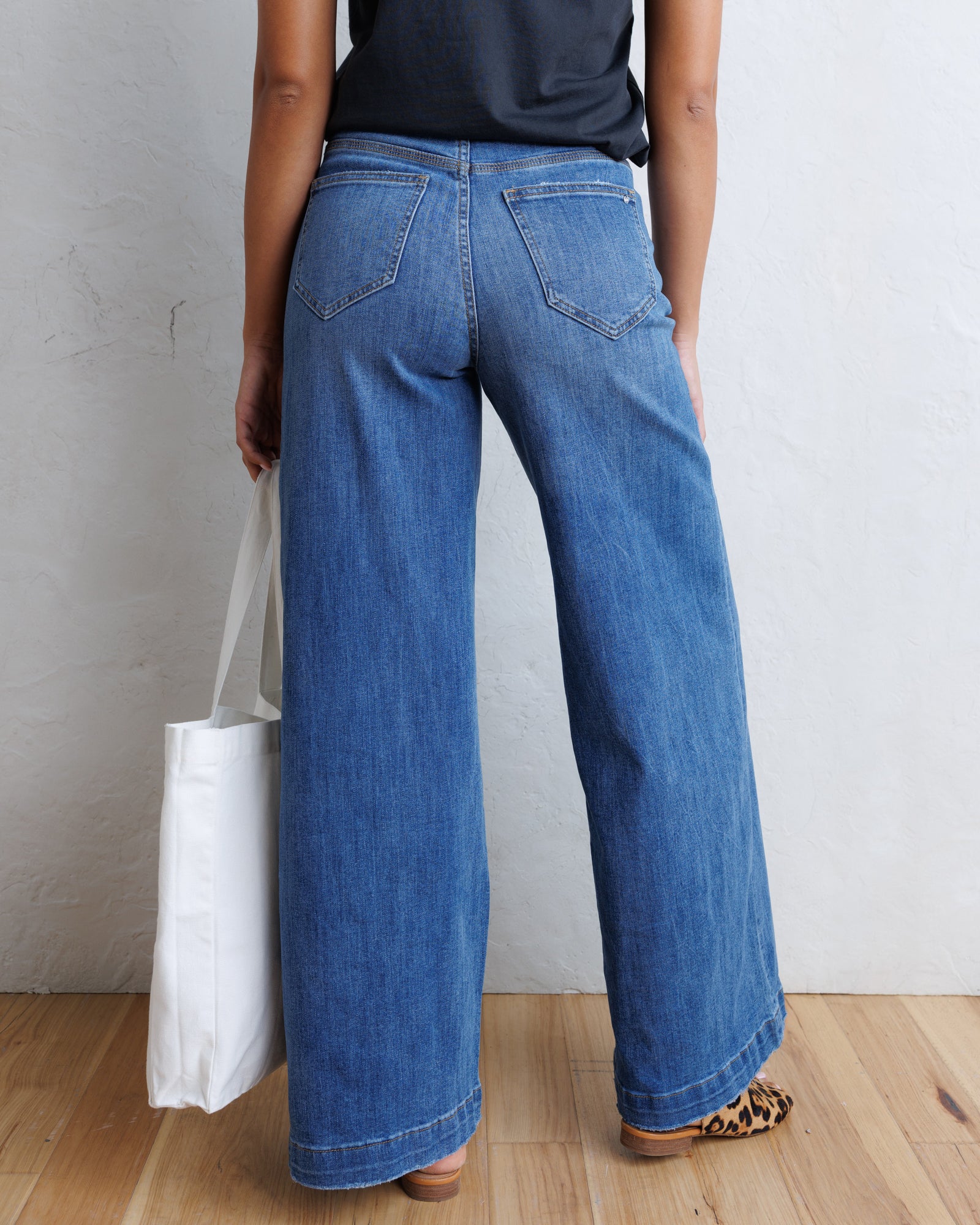 A person shown from the back wearing wide-leg blue denim jeans, a dark top, and leopard-print flat shoes, standing on a wood floor against a light textured wall and holding a white tote bag.