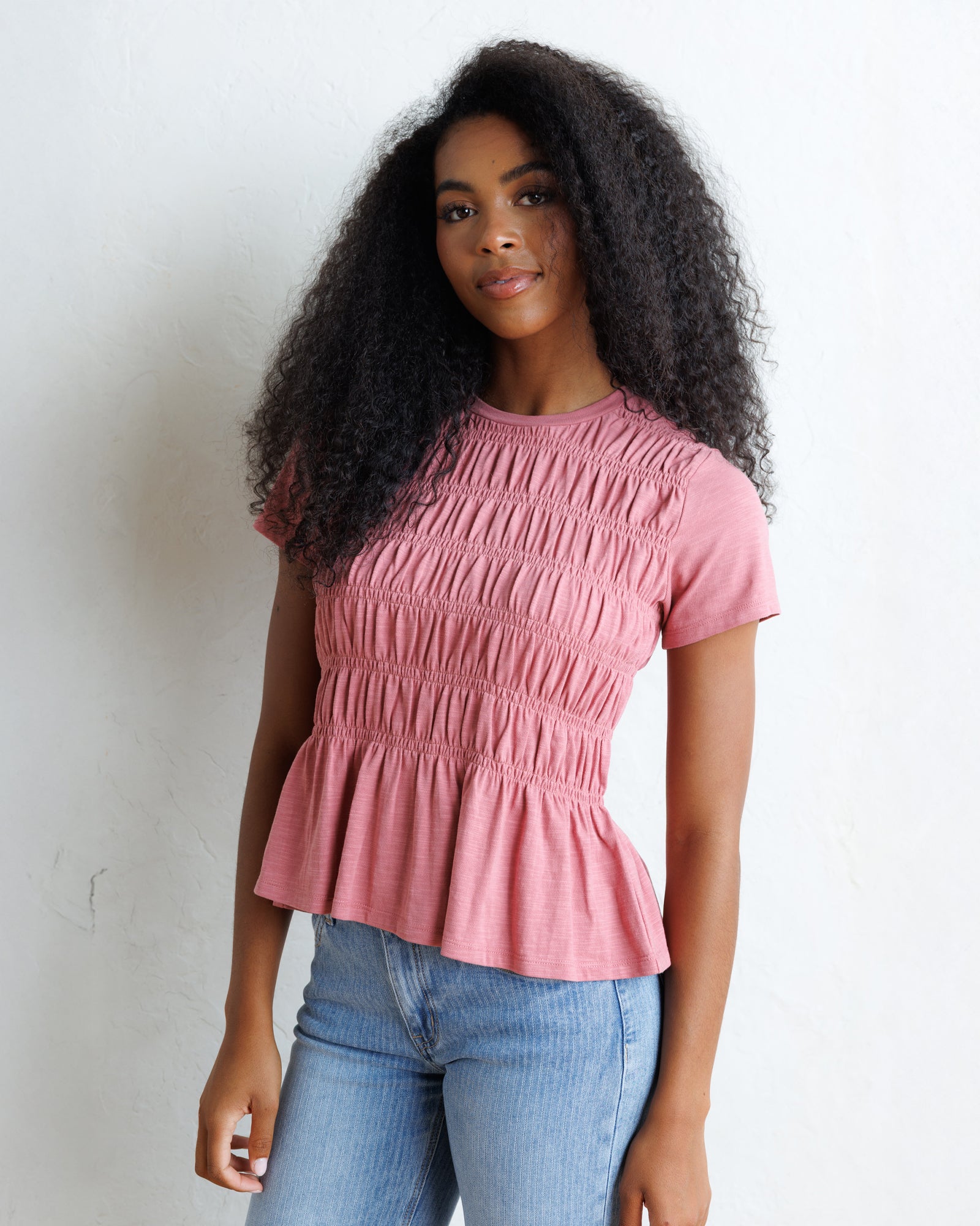 A woman with long, curly black hair wearing a dusty rose pink smocked short-sleeve top and light blue jeans, standing against a white wall.