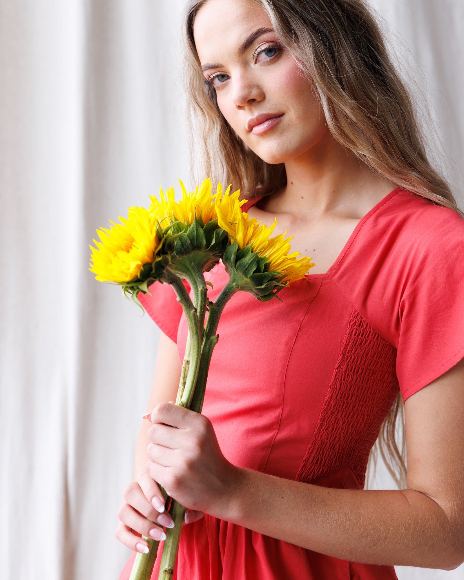 Woman in a red dress holding sunflowers against a white curtain background