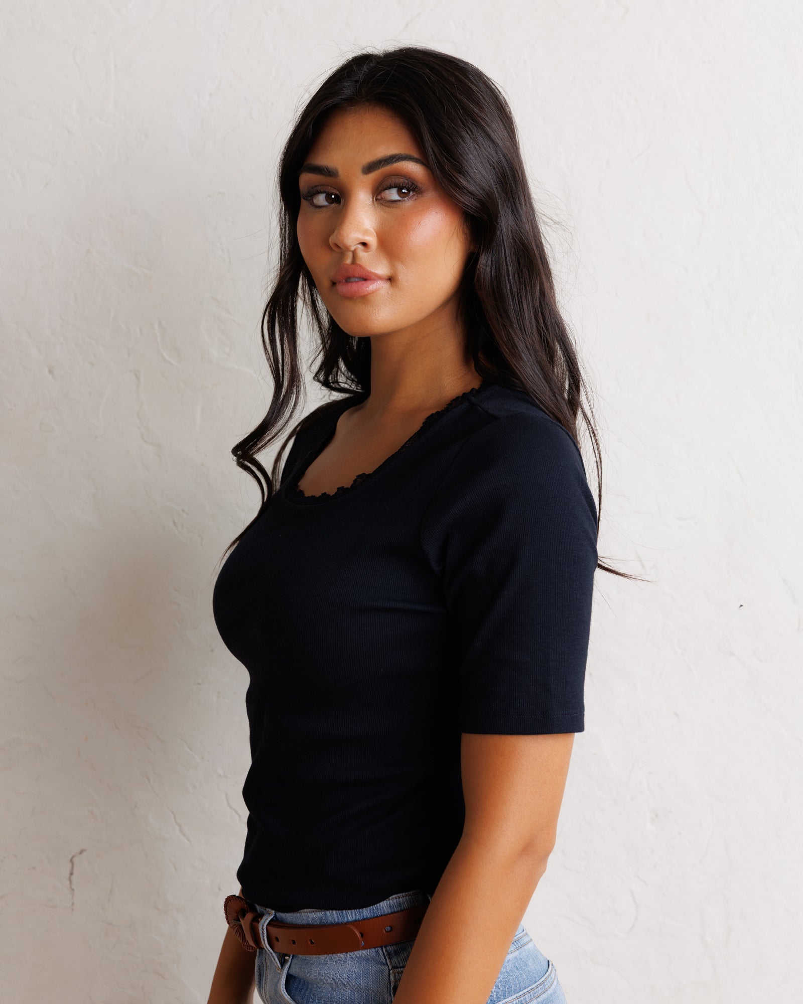 A woman with long dark hair wearing a black short-sleeve scoop-neck t-shirt and light blue jeans with a brown belt, standing against a textured off-white wall.