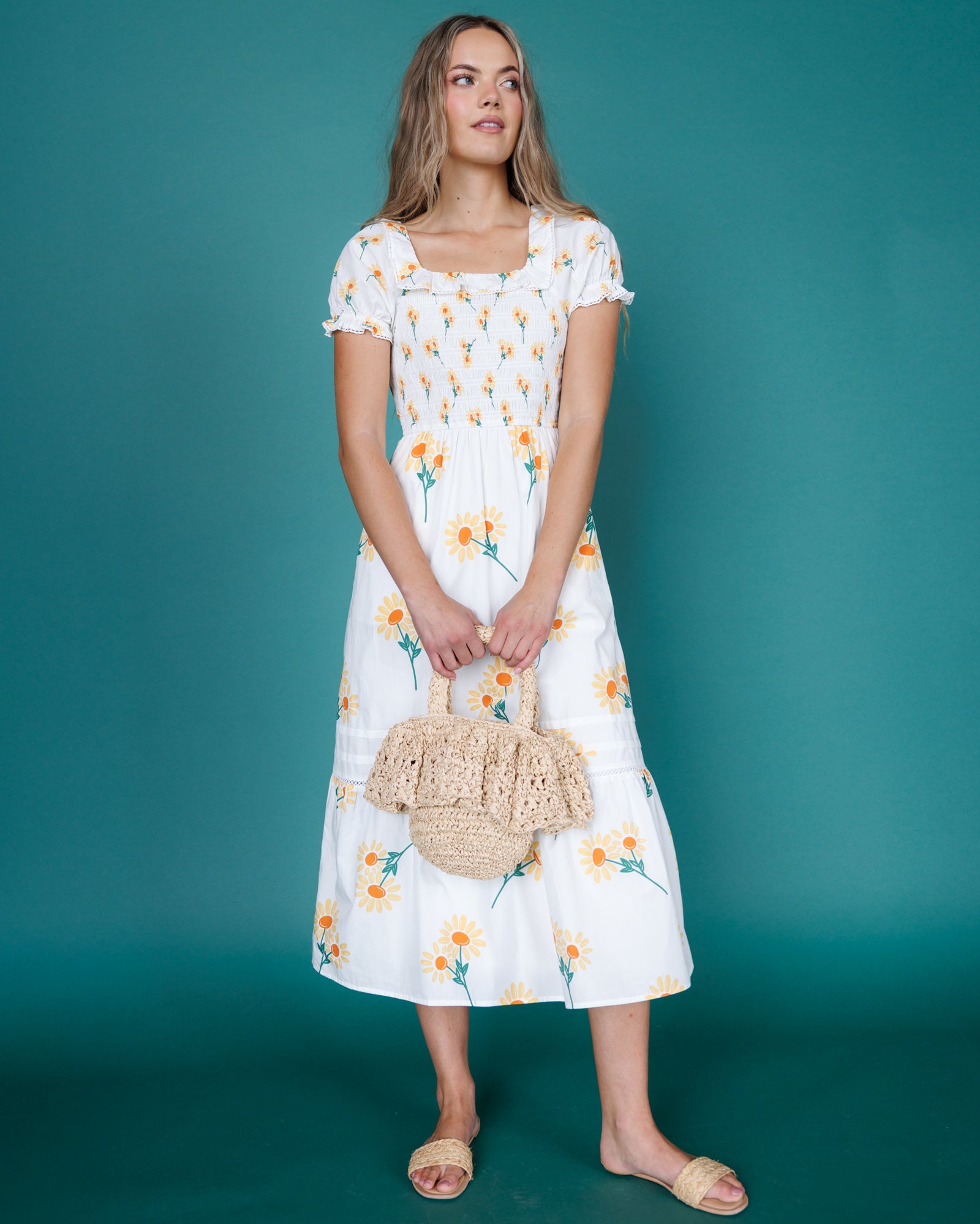 Woman wearing a white floral dress holding a woven bag against a teal background