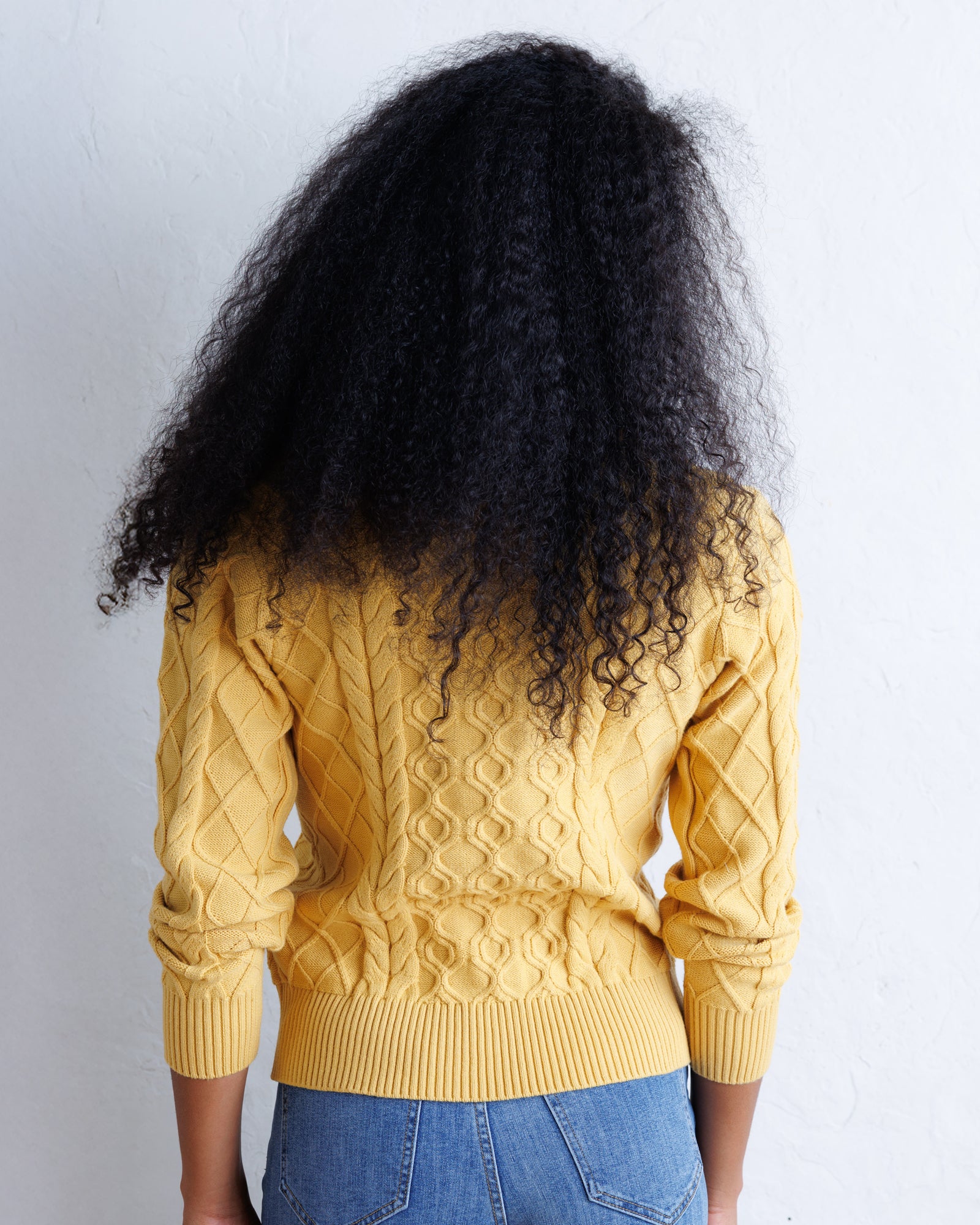 A person shown from the back wearing a mustard yellow cable-knit sweater and blue jeans, with very large dark curly hair, against a plain white wall.