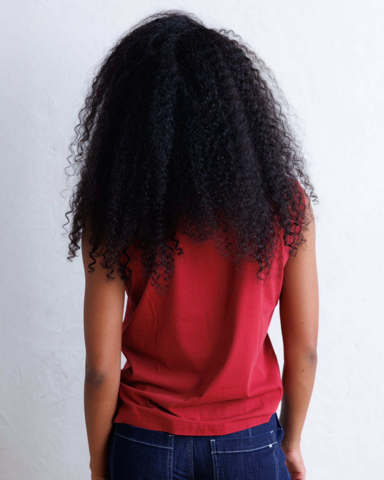 A person shown from the back wearing a red sleeveless top and dark blue jeans, with long, voluminous curly black hair, against a plain white wall.
