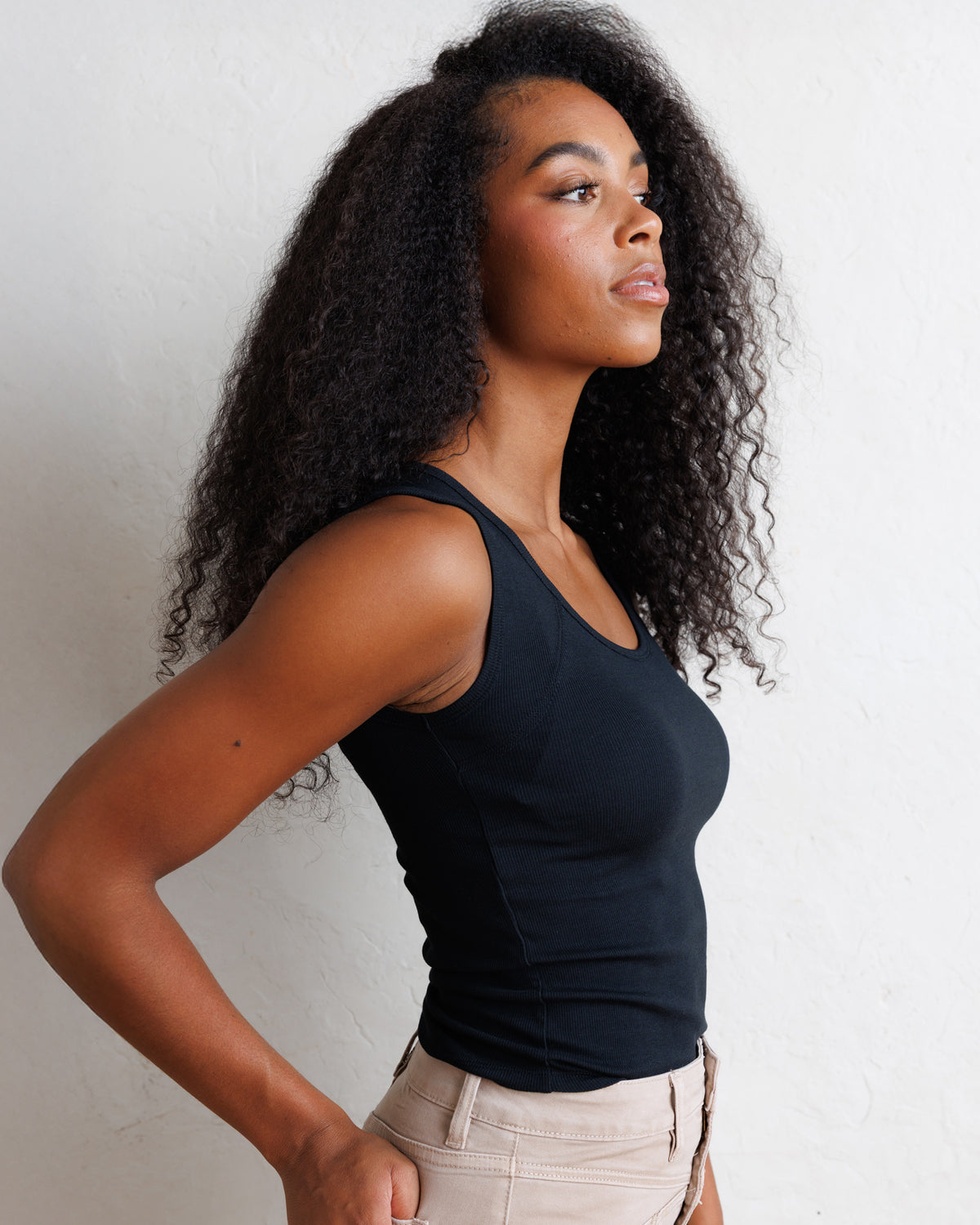 A woman with long, curly black hair is shown in profile wearing a fitted black tank top and light beige pants against a plain white wall.