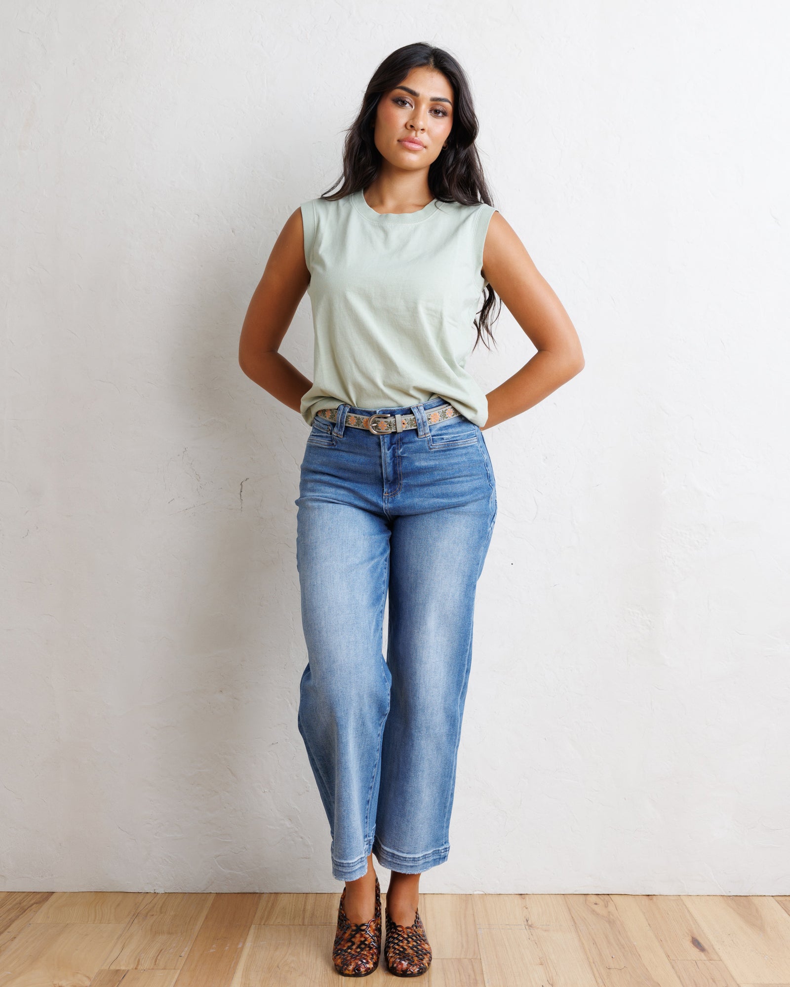 A woman stands against a textured off-white wall on a light wood floor, wearing a light sage green sleeveless top, blue cropped jeans with a gold-tone belt, and leopard-print flat shoes.