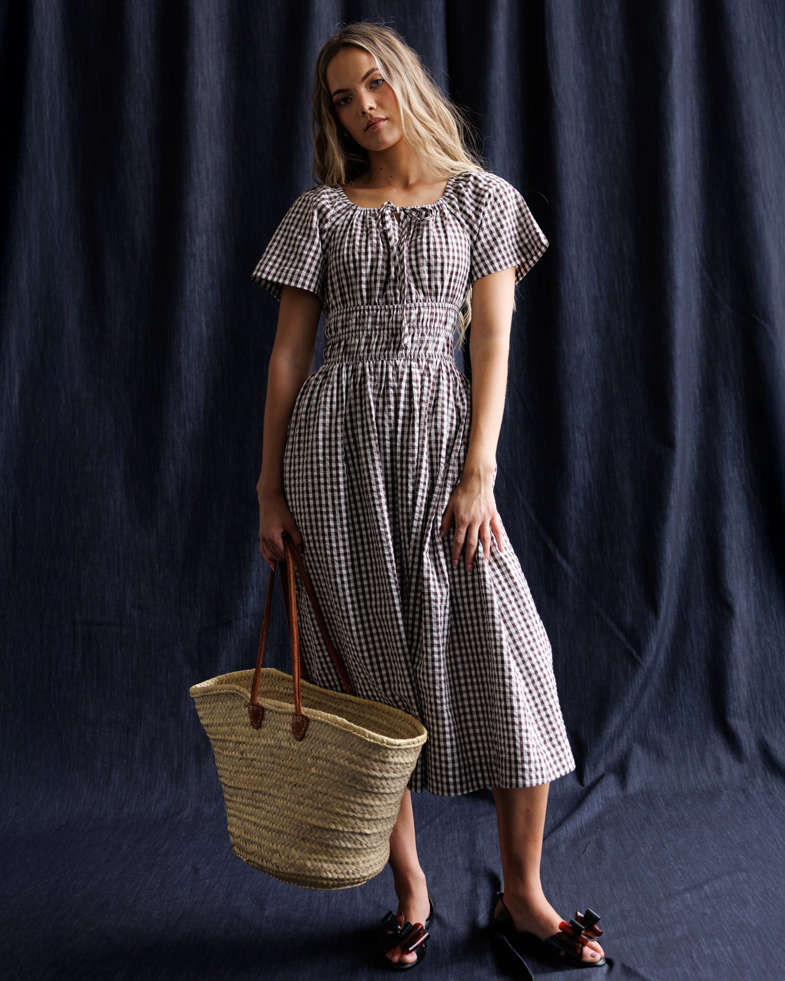 Woman in a checkered dress holding a woven bag against a dark background