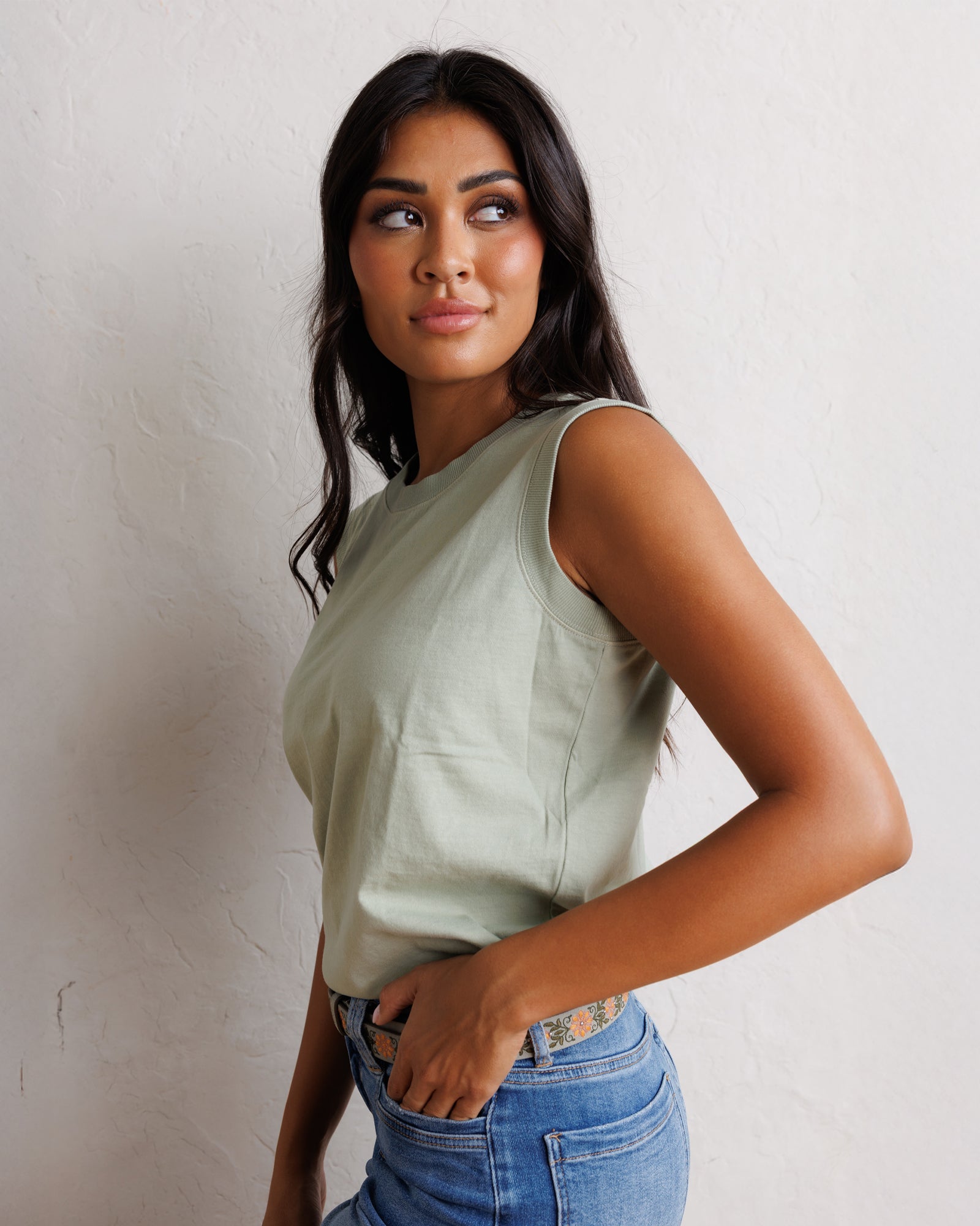 A woman wearing a sage green sleeveless crop top and blue jeans, posing against a textured off-white wall.
