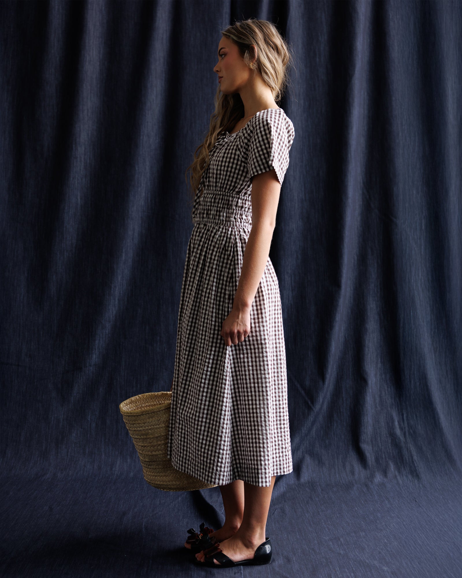 Woman wearing a checkered dress holding a woven basket against a dark background