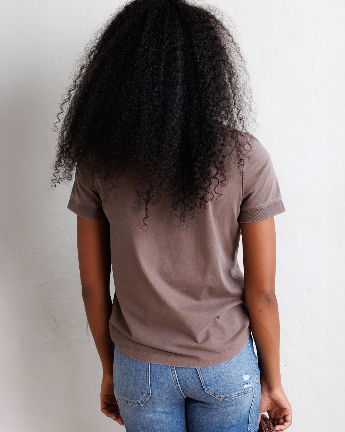 A person viewed from behind wearing a muted taupe-brown short-sleeve t-shirt and blue jeans, standing against a plain off-white wall.