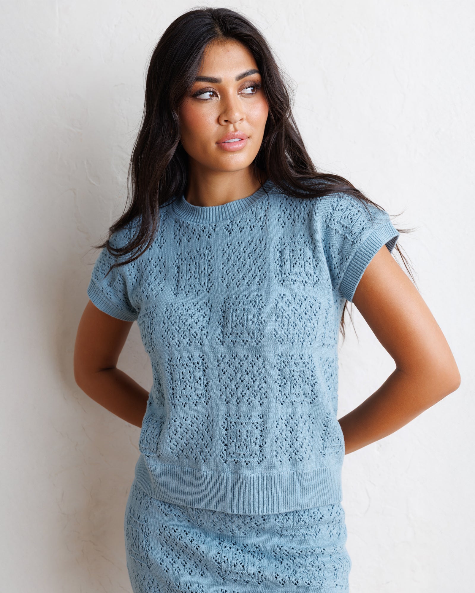 A woman wearing a light blue short-sleeve knit sweater with an openwork textured pattern, standing against a textured off-white wall.