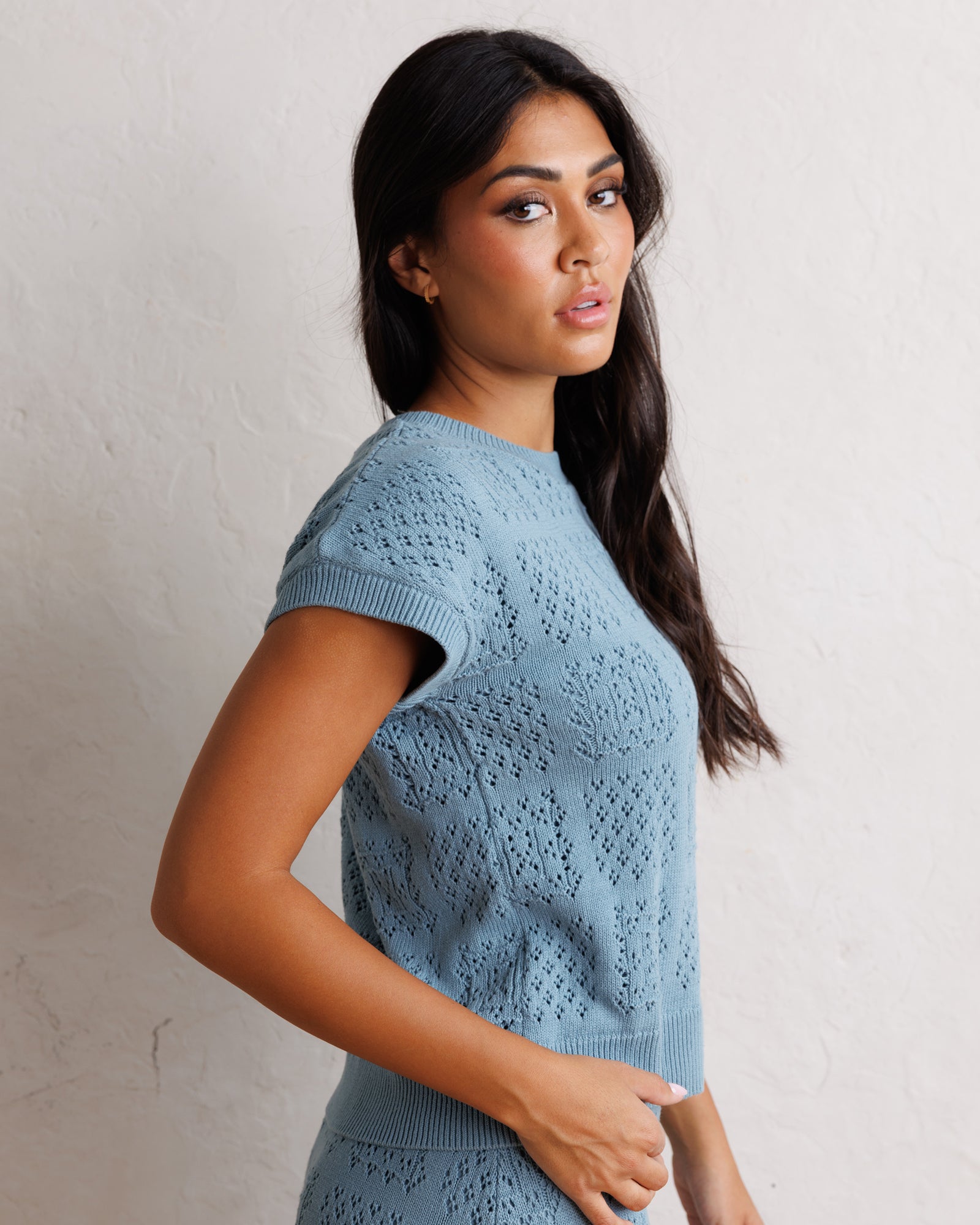 A woman wearing a light blue short-sleeve knit sweater with an openwork textured pattern, standing against a textured off-white wall.