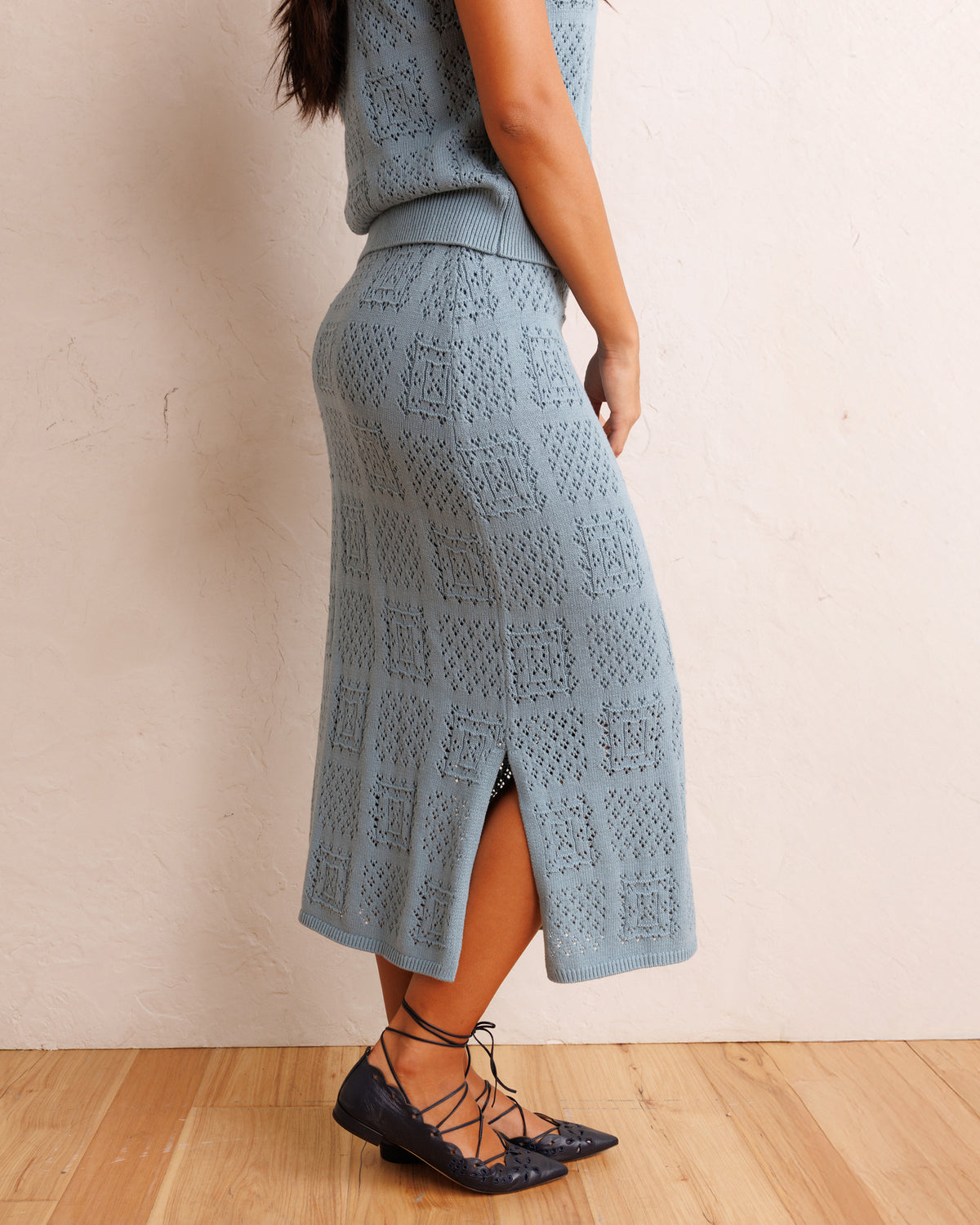 A woman wearing a light blue open-knit midi skirt with a side slit, paired with a matching top and dark lace-up flats, standing against a textured beige wall on a wood floor.