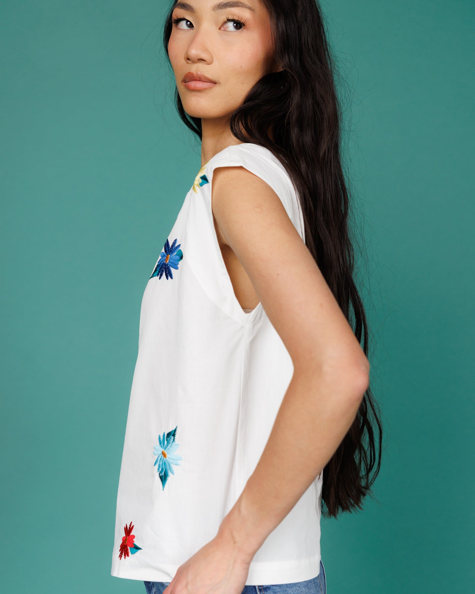 Woman wearing a white sleeveless top with colorful floral embroidery against a teal background