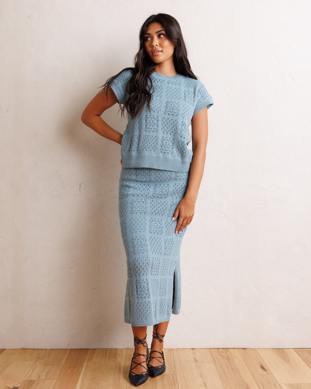A woman models a light blue knit two-piece set consisting of a short-sleeve top and a matching midi skirt with a front slit, set against a neutral beige wall and light wood floor.