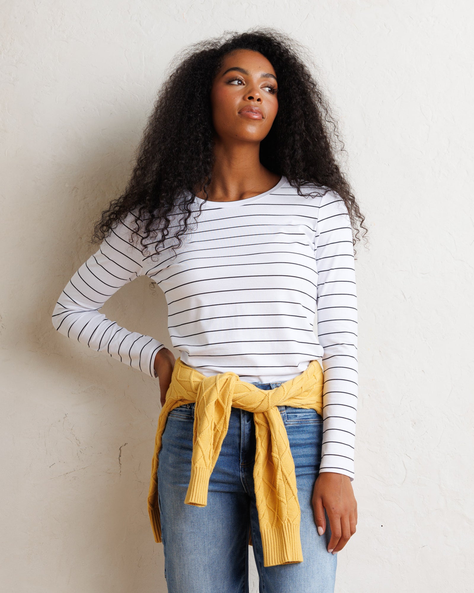 A woman with long curly black hair stands against a textured off-white wall, wearing a white long-sleeve crewneck top with thin black horizontal stripes, blue jeans, and a yellow knit sweater tied around her waist.