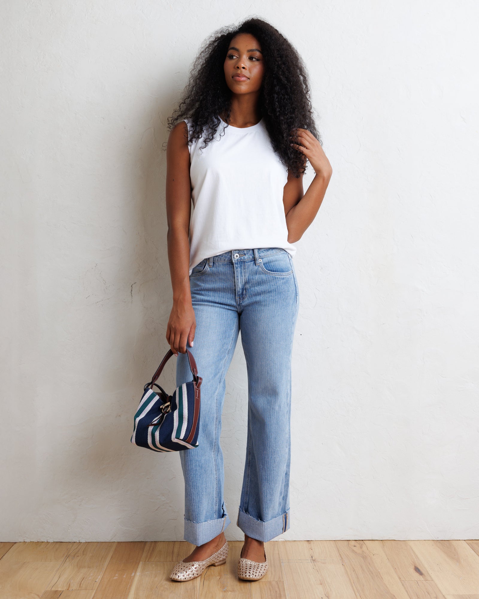 A woman stands against a textured off-white wall on a light wood floor, wearing a white sleeveless top, light-wash blue jeans with rolled cuffs, and beige flat shoes. She holds a small striped handbag.