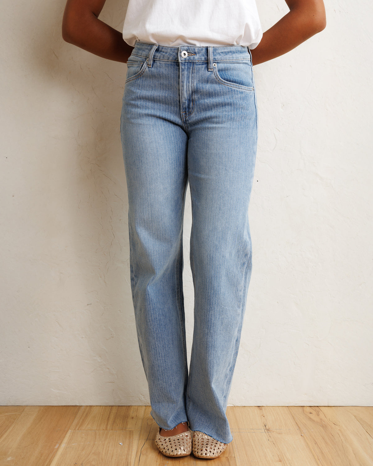 A person wearing light-wash blue wide-leg jeans, a white top, and beige woven flat shoes, standing against a textured off-white wall on a light wood floor.