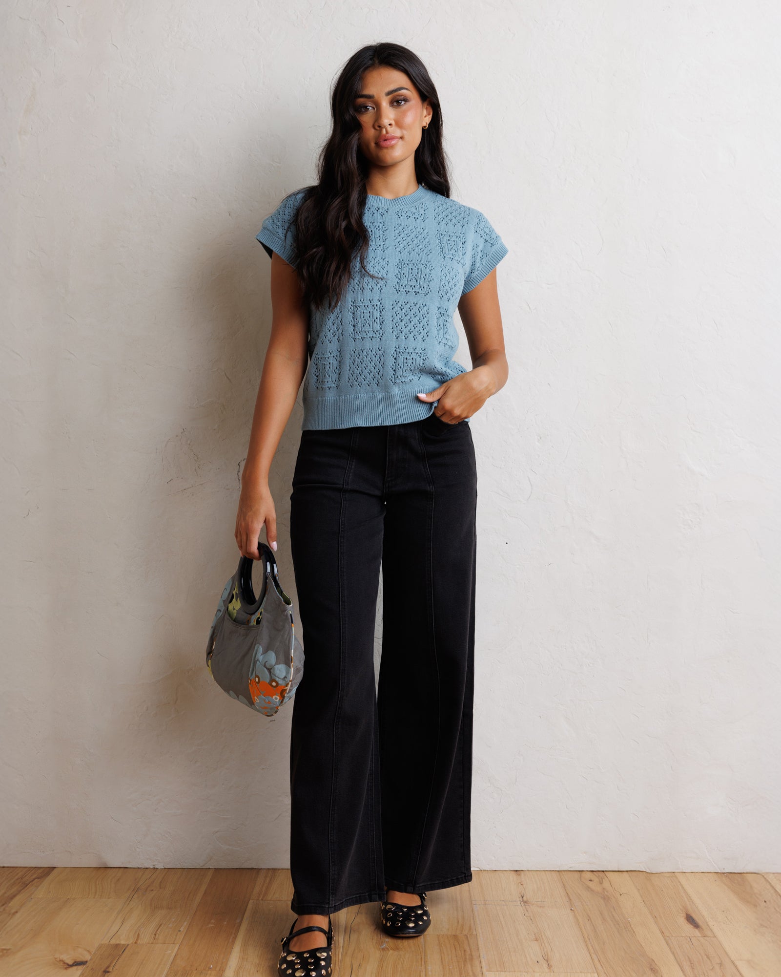 A woman stands against a textured off-white wall on a light wood floor, wearing a light blue short-sleeve knit top with a geometric pattern, black wide-leg pants, and black polka-dot flat shoes, while holding a gray patterned handbag.