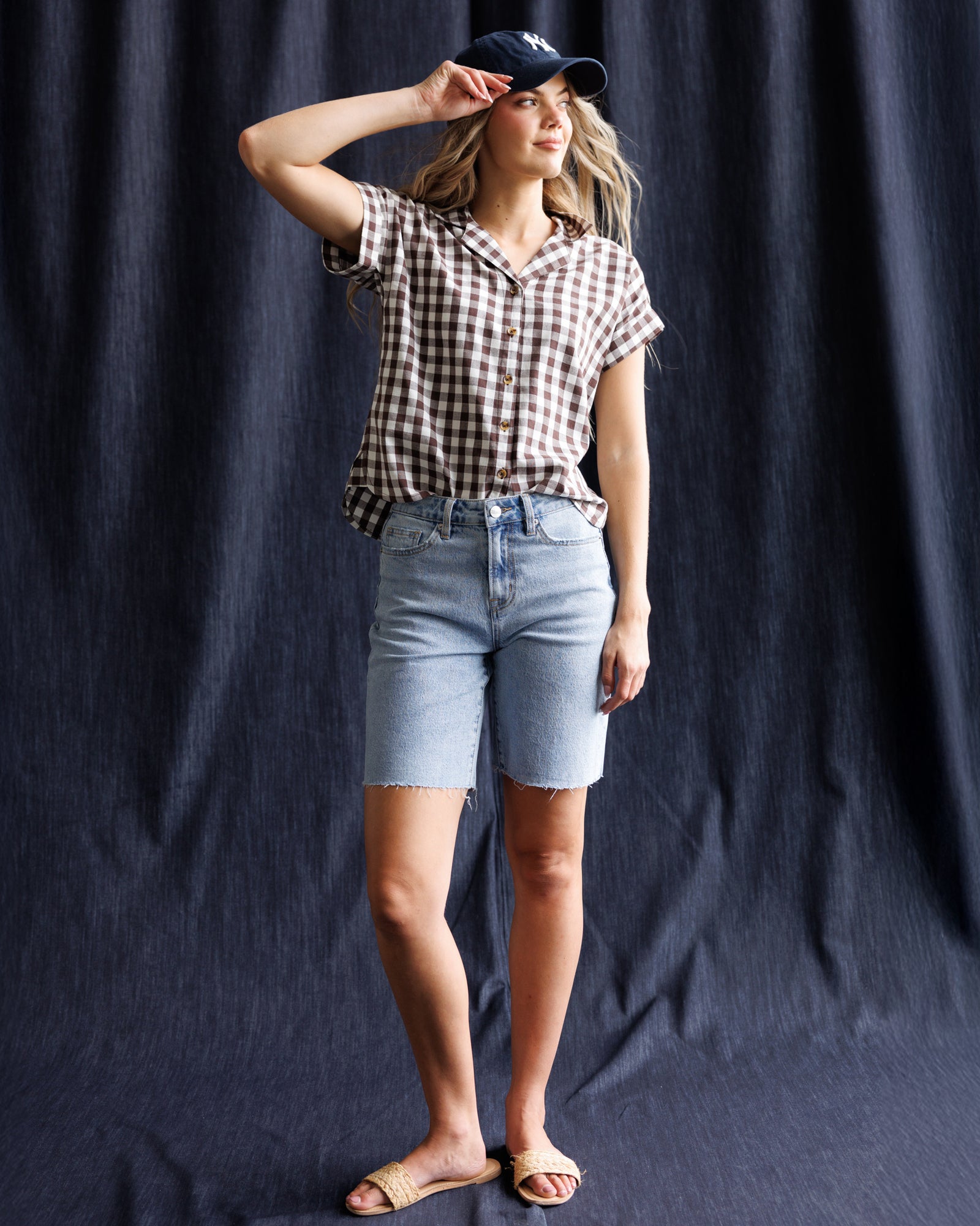 Woman wearing a checkered shirt, denim shorts, and a cap against a dark background