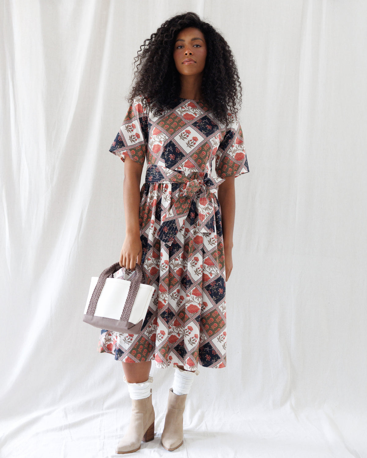 Front view of a patchwork floral midi dress featuring short sleeves, a crew neckline, and a gathered A-line skirt cinched with a matching tie belt at the waist.