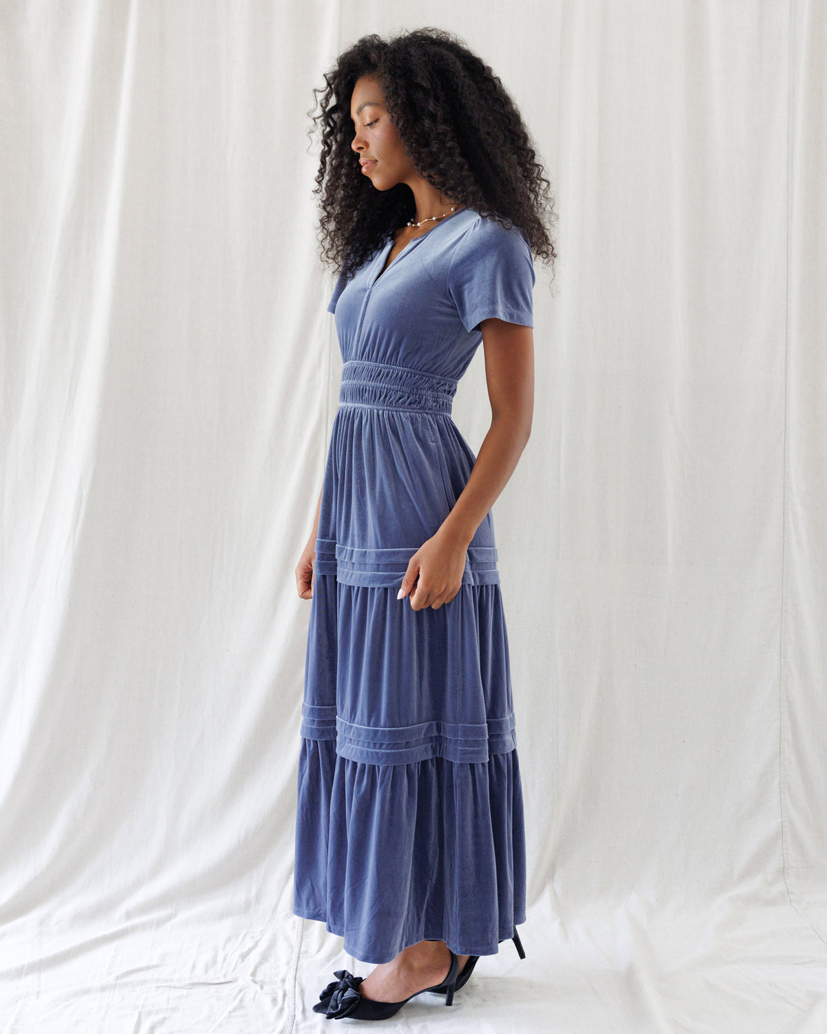 Side profile showing the defined high-waist smocking, classic short-sleeve cut, and sweeping tiered A-line silhouette of the soft blue velvet dress.