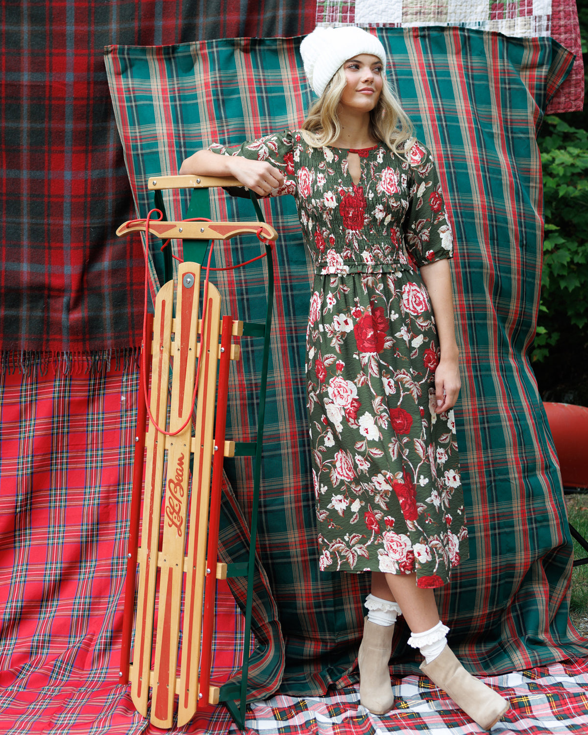 A woman wearing a green floral midi dress, white knit beanie, white socks, and beige ankle boots stands beside a wooden sled in front of red and green plaid fabric backdrops.