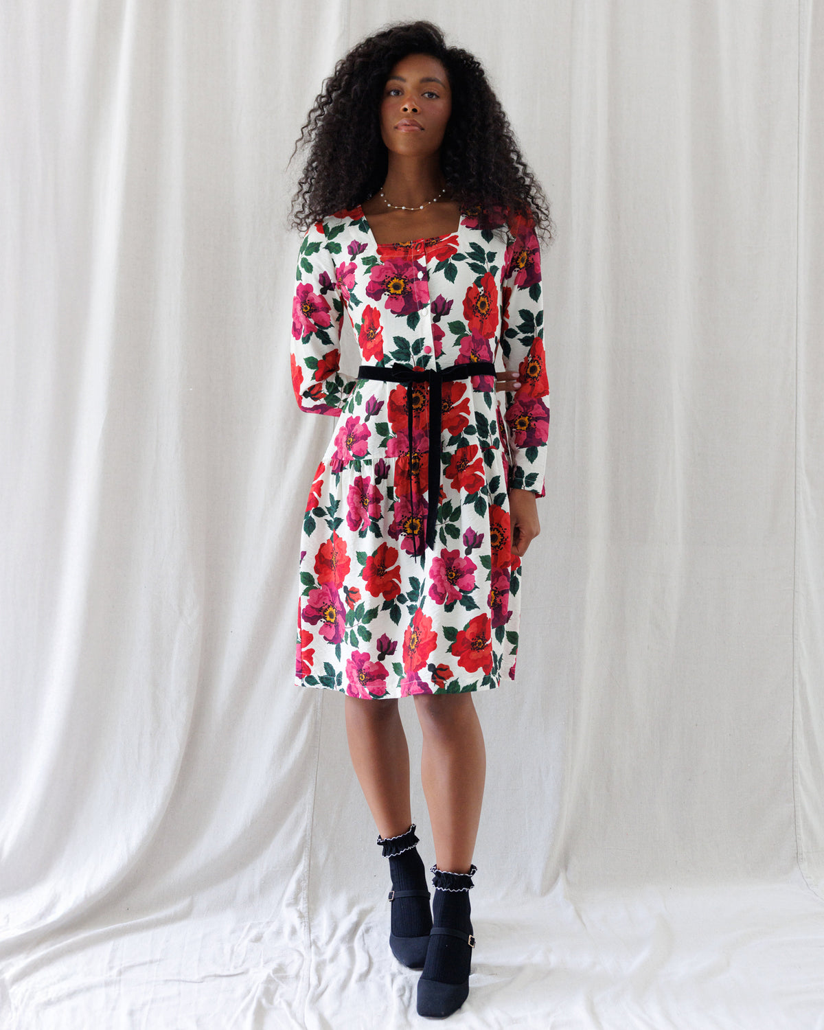A woman modeling a white long-sleeve knee-length dress with a vibrant red and pink floral print and a black waist tie, paired with black ankle boots, set against a draped white fabric backdrop.