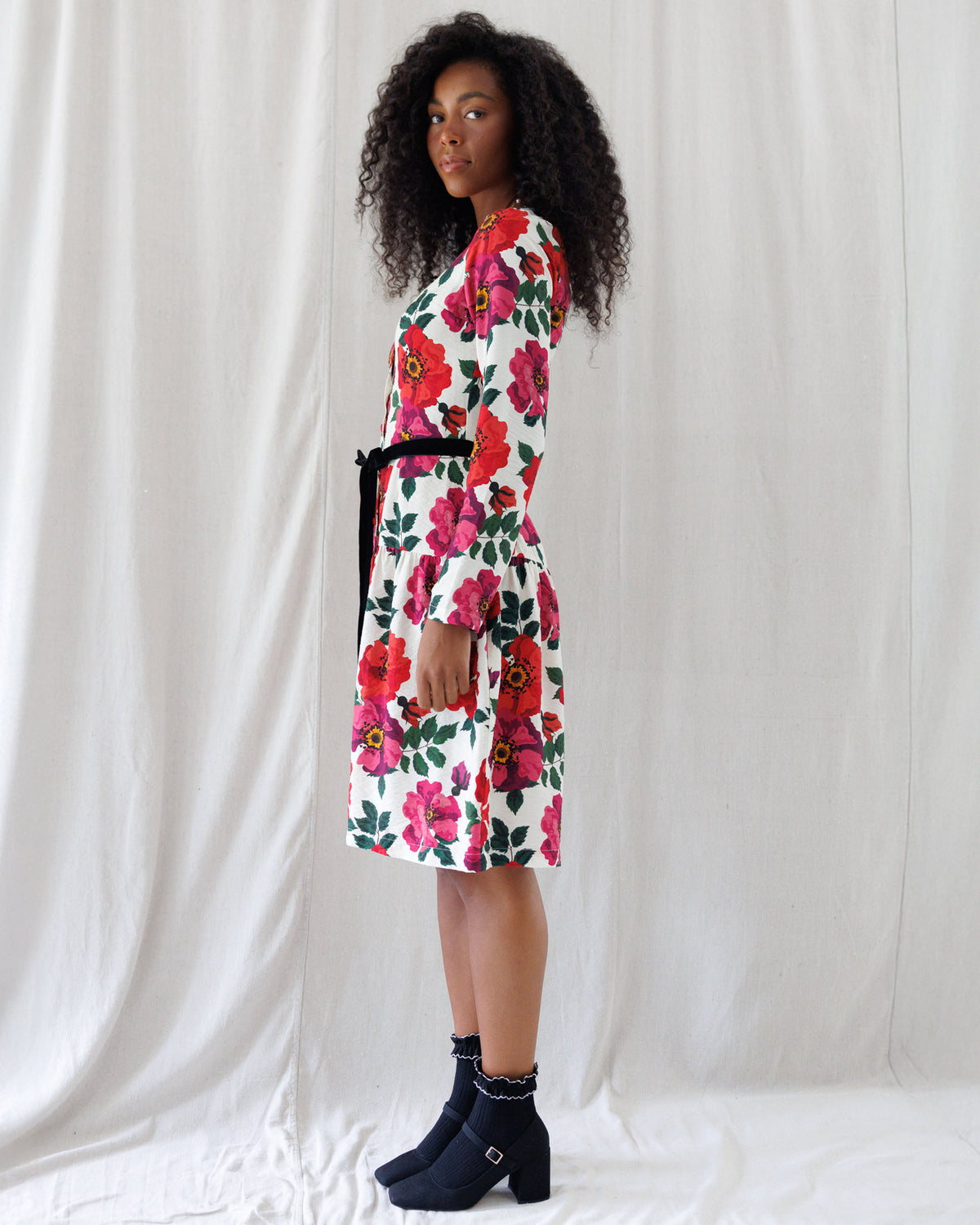 A woman stands in profile wearing a knee-length white dress with a bold red, pink, and green floral print and a black waist tie, paired with dark ankle boots, against a draped white fabric backdrop.