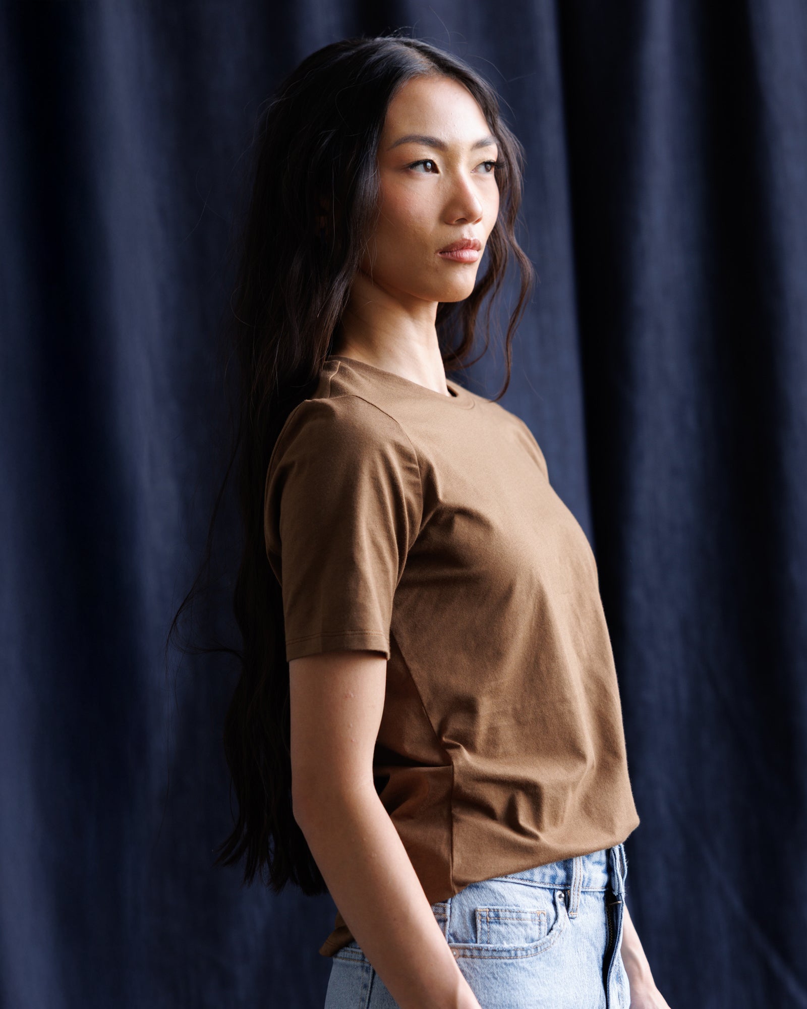 A woman with long dark hair is shown in profile wearing a brown short-sleeve crewneck t-shirt and light blue jeans, standing against a dark navy blue curtain backdrop.