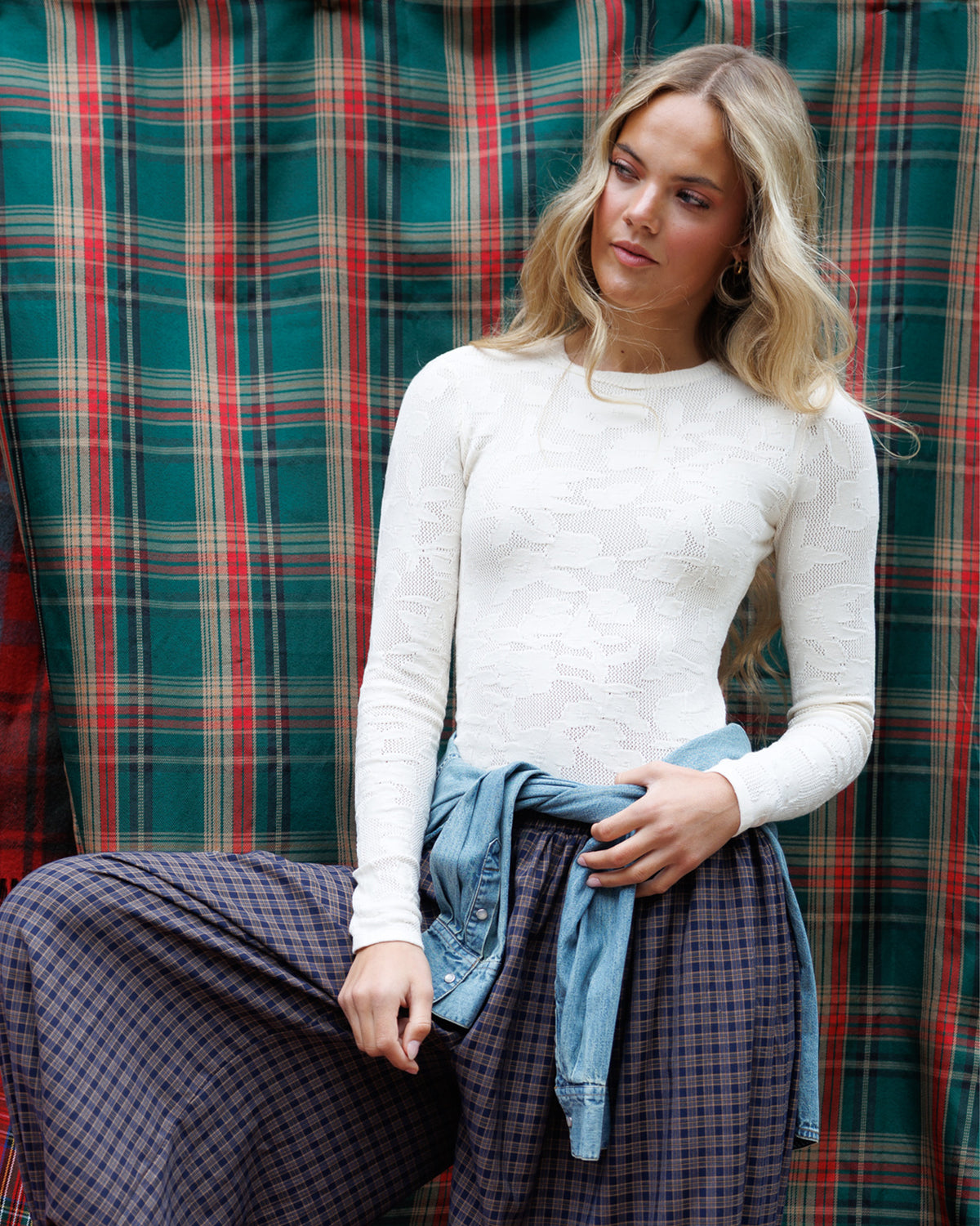 A woman wearing a fitted white textured long-sleeve top and dark plaid wide-leg pants, with a light blue denim shirt tied around her waist, posed in front of a green, red, and beige plaid fabric backdrop.