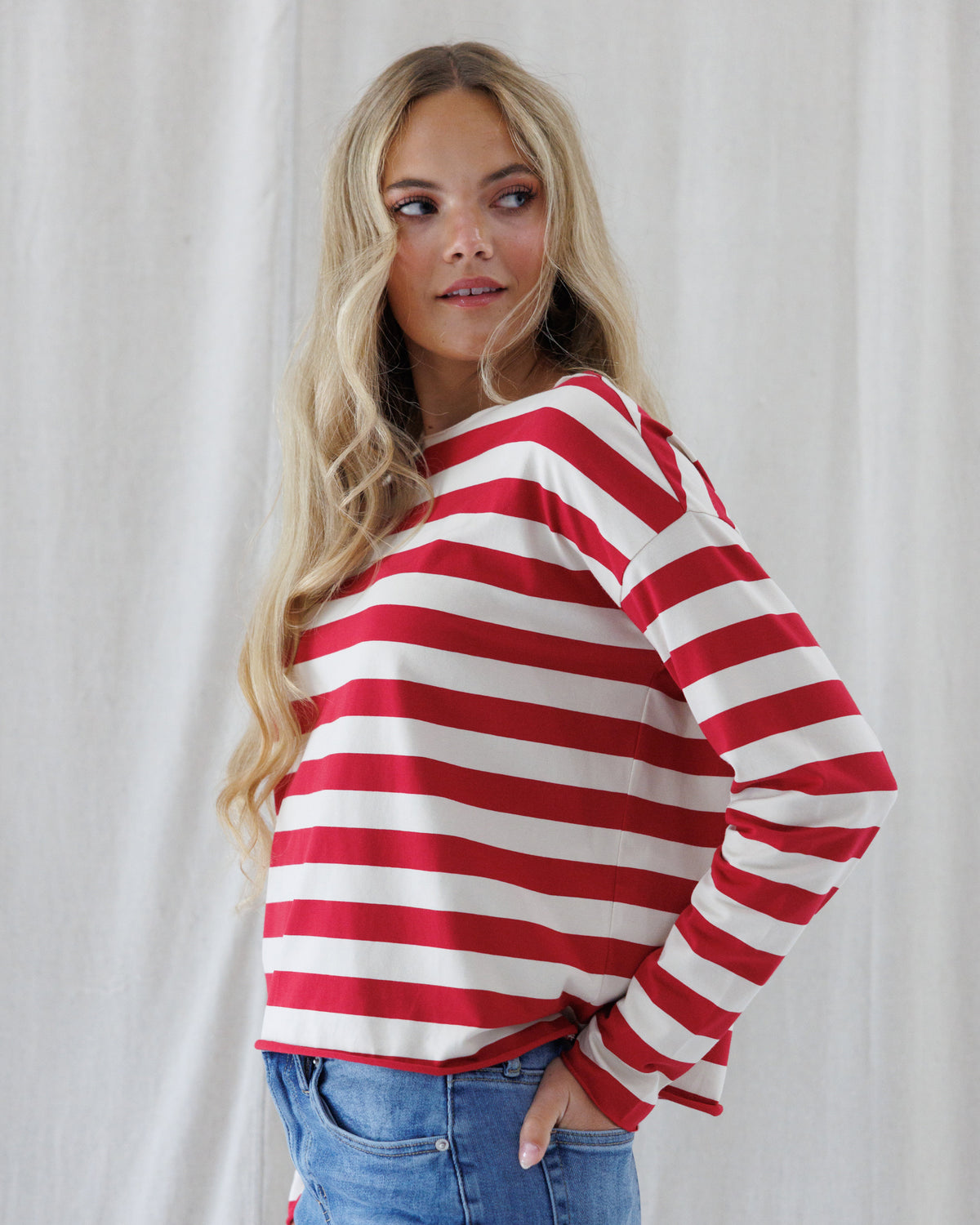 A woman wearing a red and off-white horizontally striped long-sleeve top with a wide boat neckline, paired with blue jeans, standing against a light neutral curtain background.