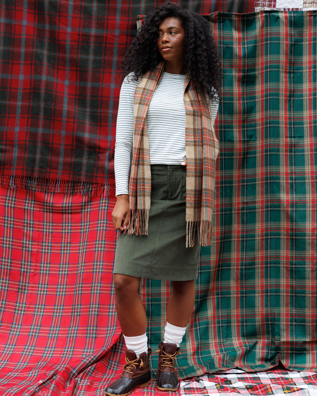 A woman stands in front of red and green plaid fabric backdrops, wearing a white striped long-sleeve top, an olive green skirt, a beige and brown plaid scarf, white socks, and brown lace-up boots.