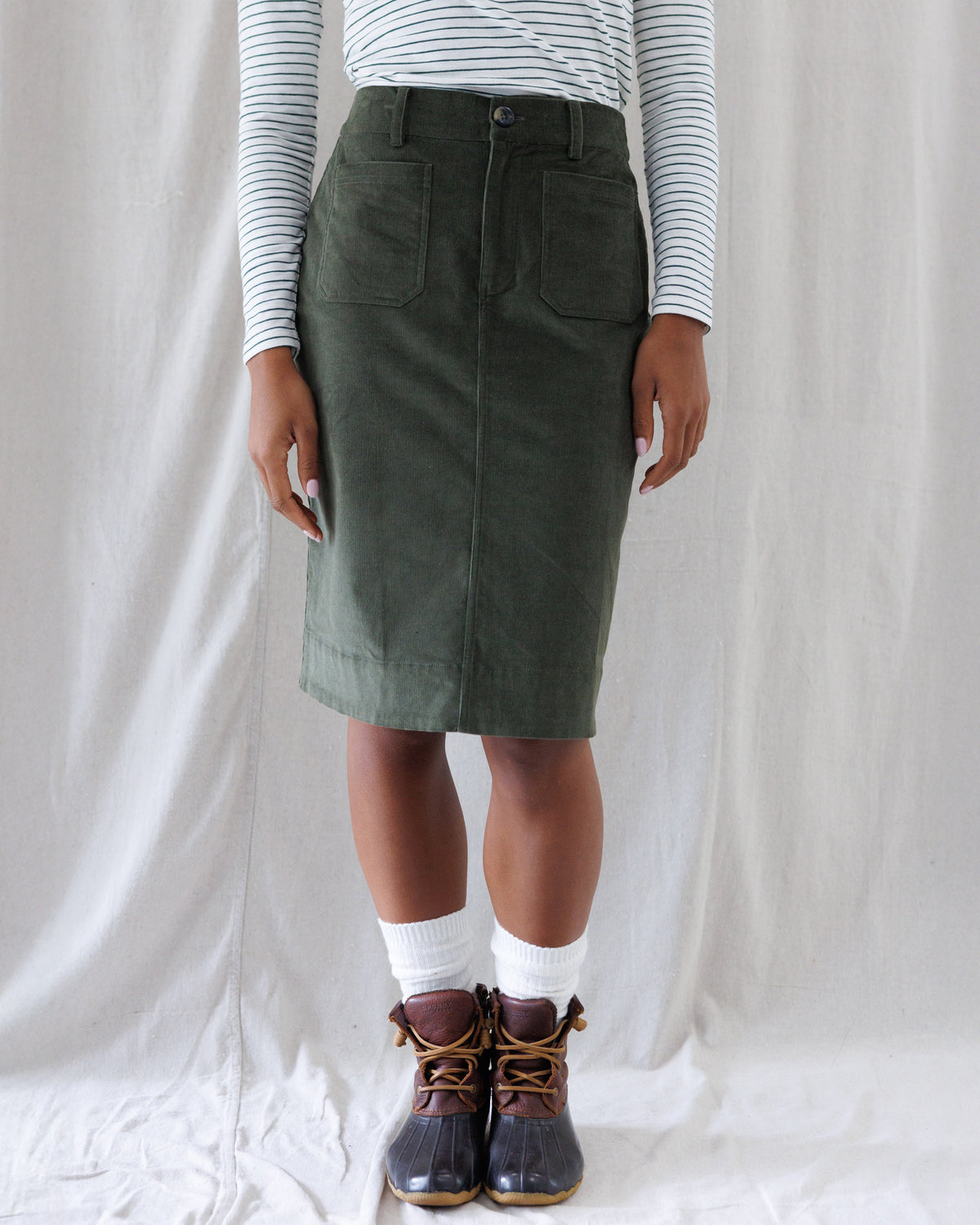 Front view of an olive green corduroy knee-length skirt featuring a high-rise waist, button fly, belt loops, and deep front patch pockets