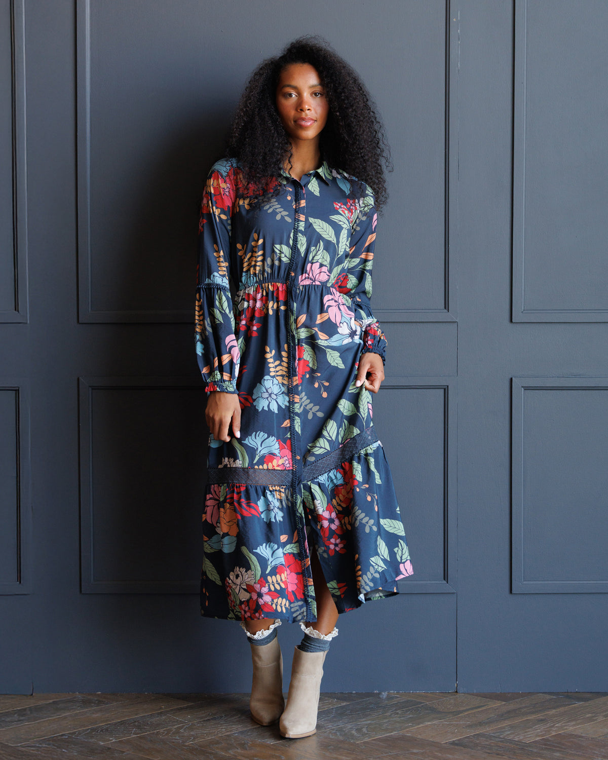 A woman with curly black hair stands against a dark blue paneled wall wearing a long-sleeved navy blue midi dress with a colorful floral print, paired with beige ankle boots.