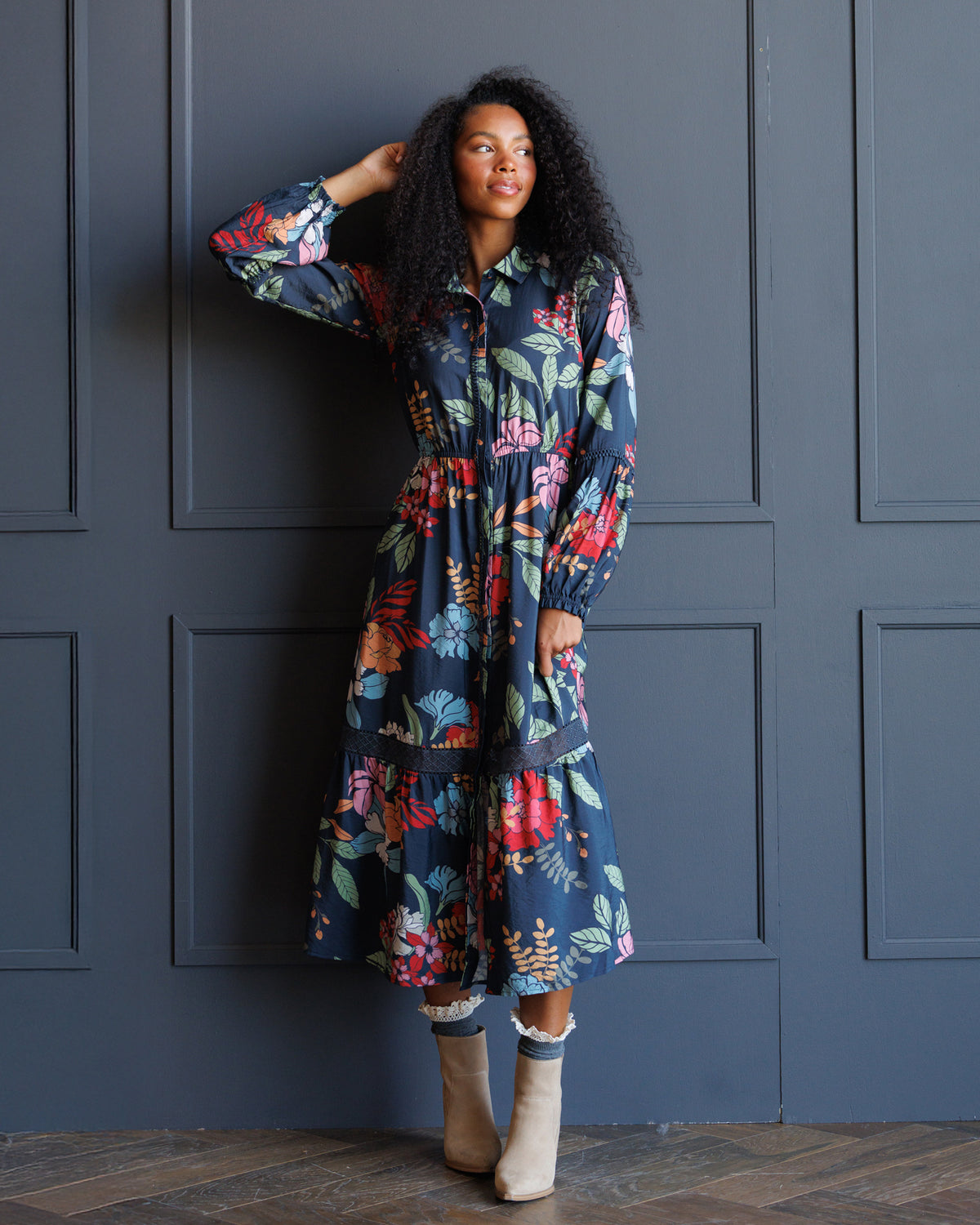 A woman with curly black hair models a long-sleeved midi dress with a colorful floral print on a dark navy background. She is standing in front of a dark gray paneled wall, wearing beige ankle boots.