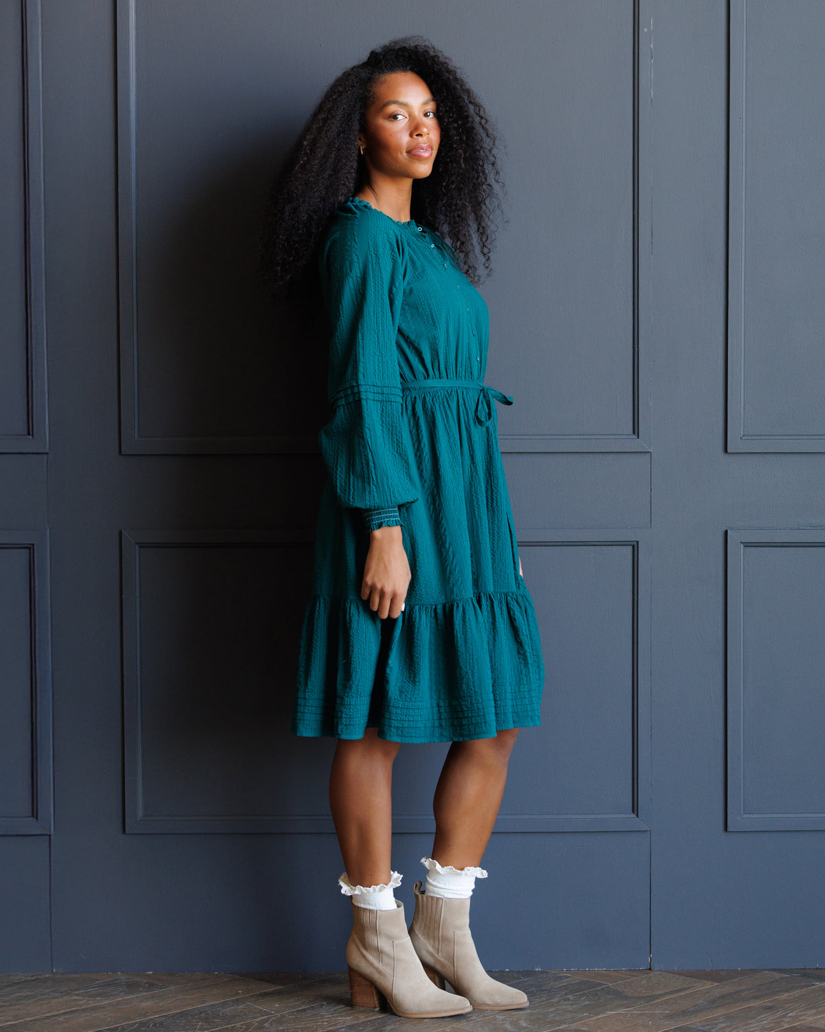 A woman wearing a teal green tiered midi dress with long puff sleeves and a tie waist, paired with beige ankle boots and white ruffled socks, posing against a dark blue paneled wall.