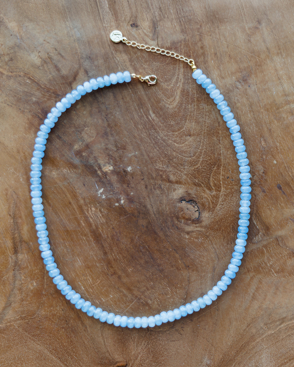 A light blue beaded necklace with a gold chain extender and lobster clasp, displayed on a rustic wooden surface.