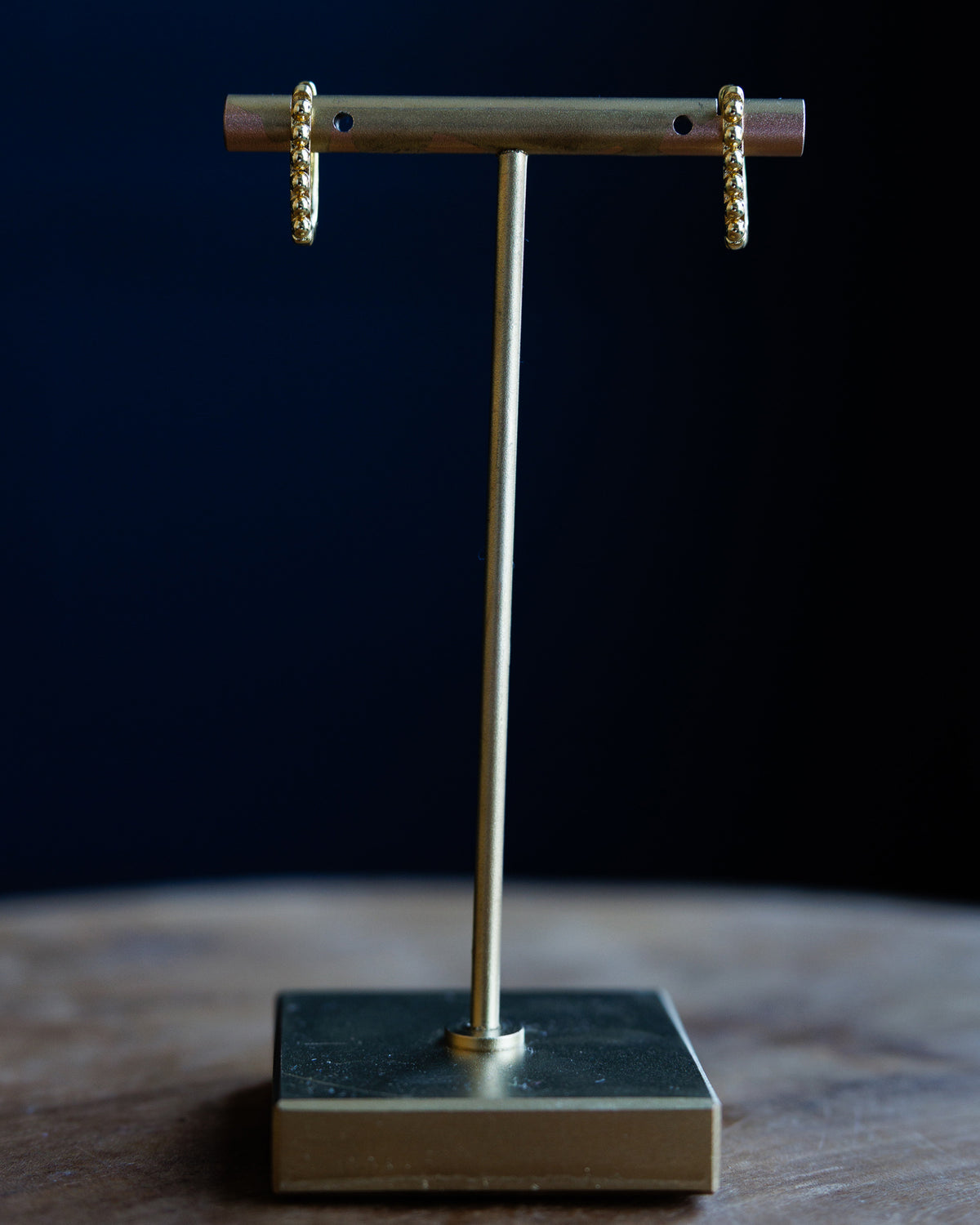 A pair of gold beaded drop earrings displayed on a brass T-stand against a dark navy blue background.