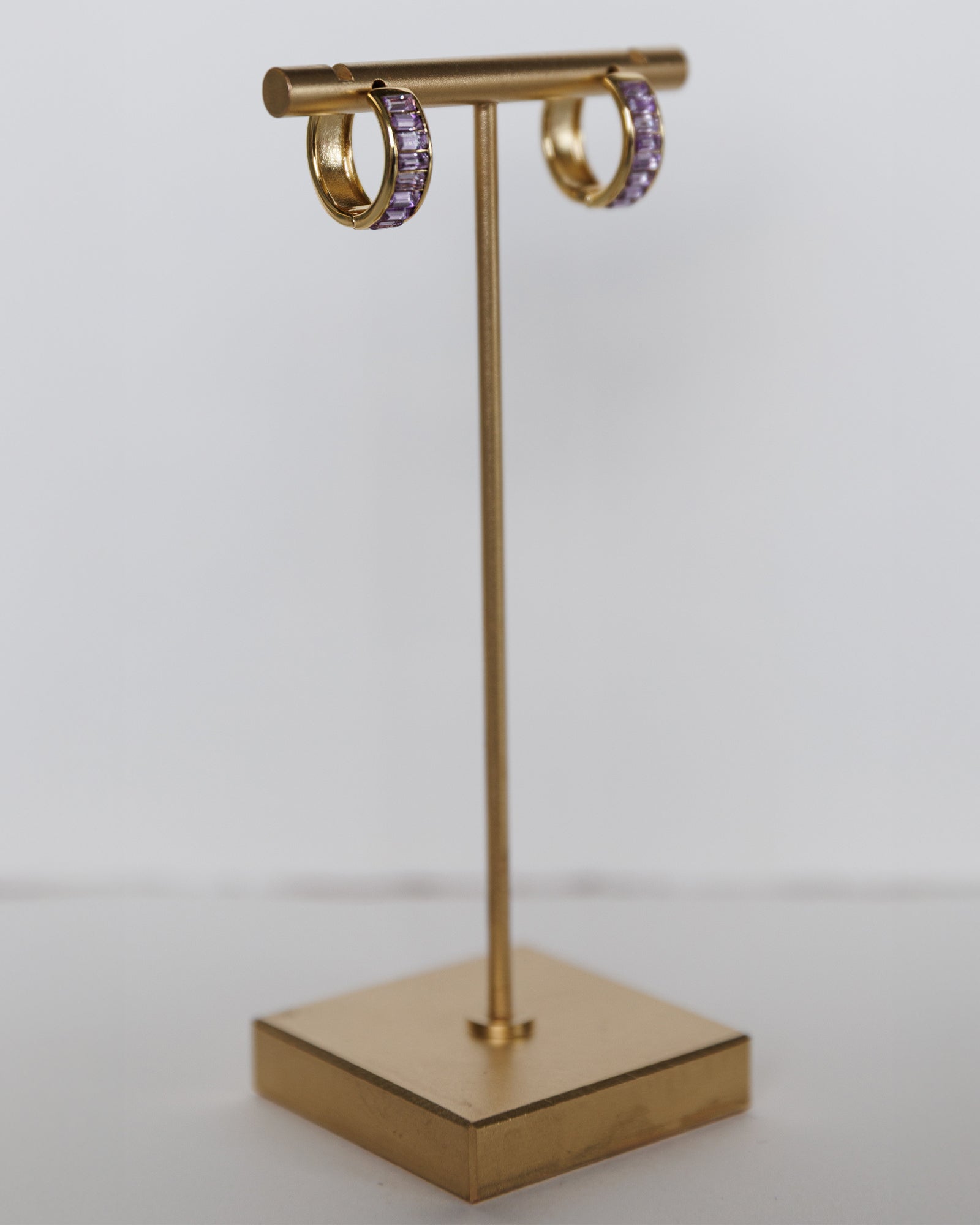 Gold earring stand with purple gemstone hoop earrings on a white background