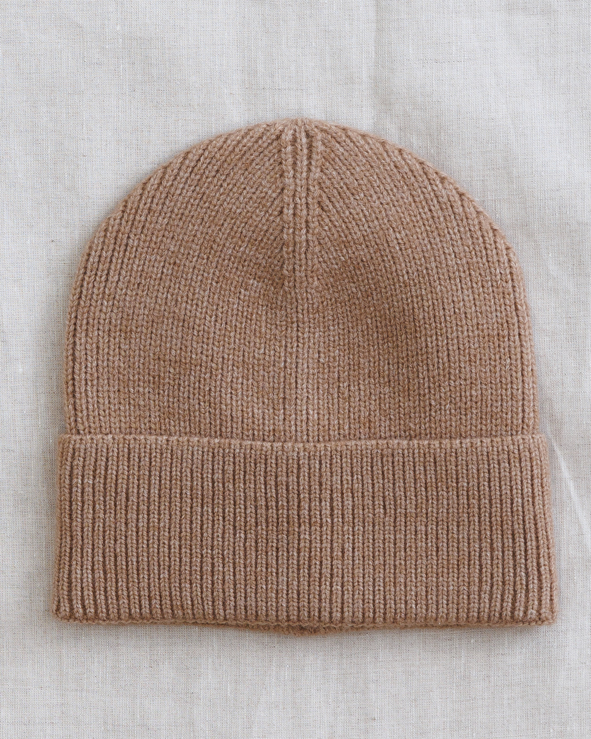 A light brown ribbed knit beanie hat laid flat on a textured off-white linen background.